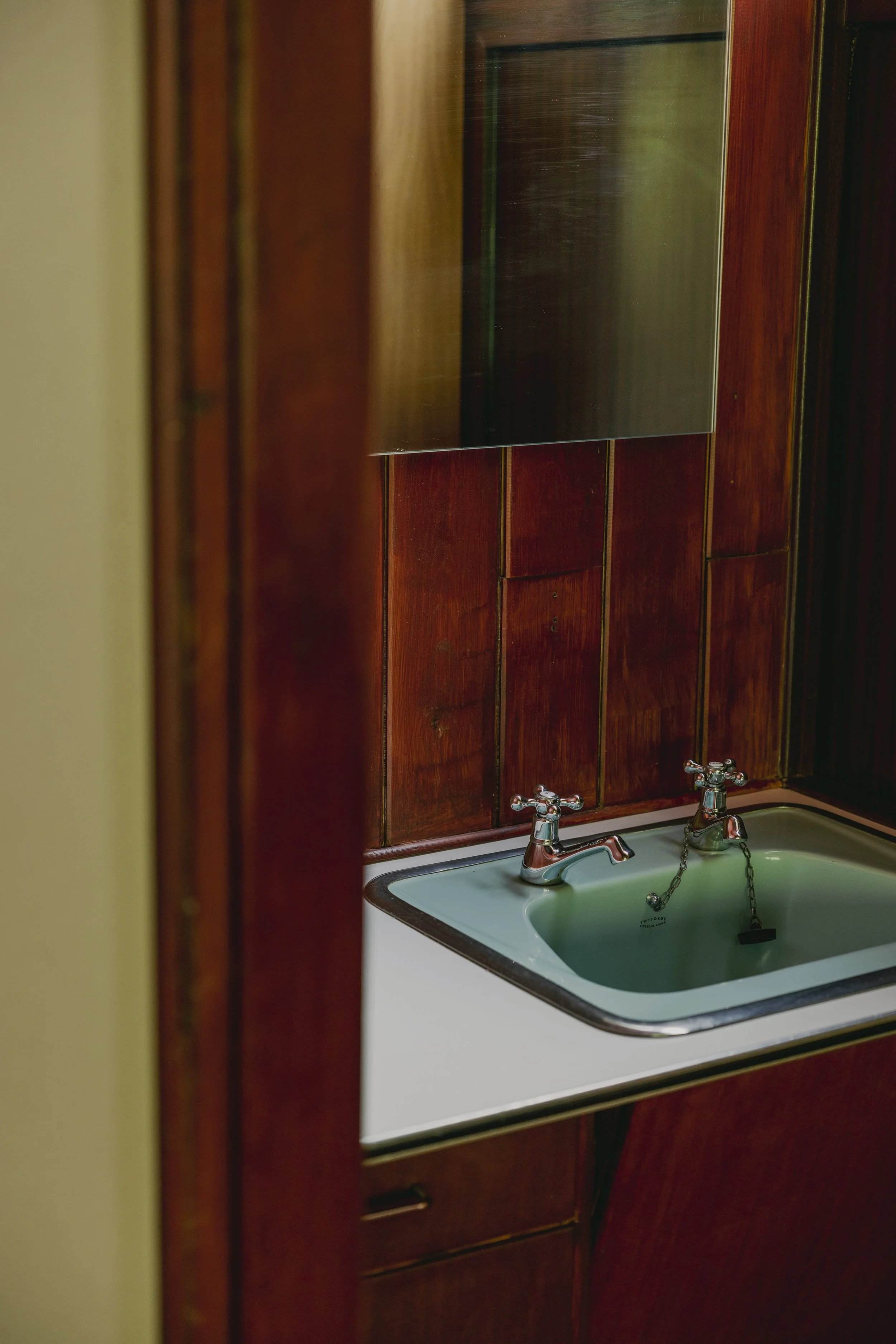 A vintage bathroom sink with a large mirror above it, set against dark wood paneling.