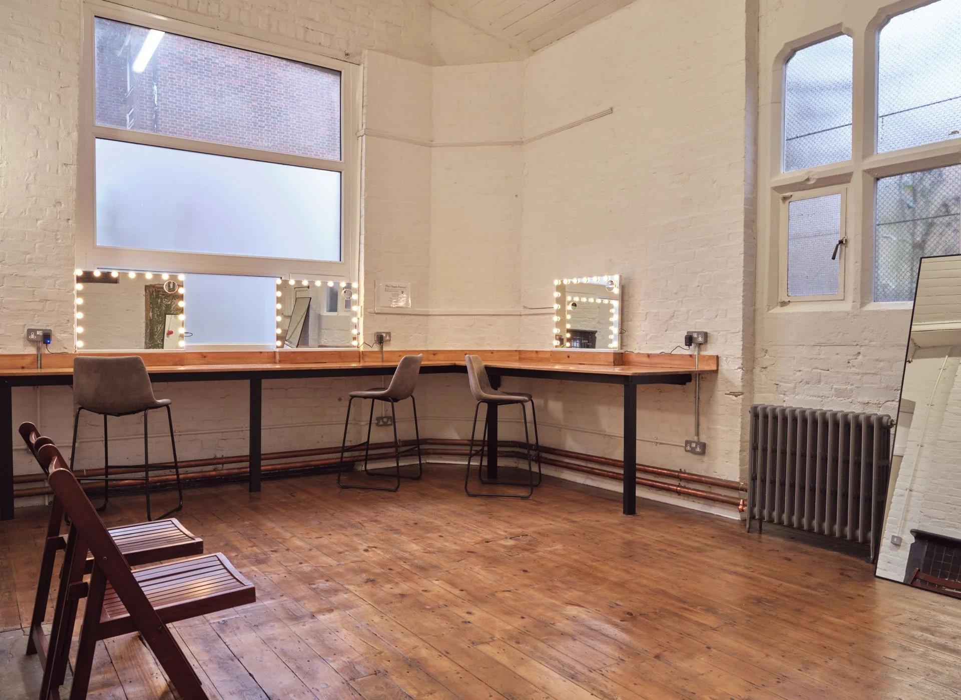 A dressing room or makeup area with white brick walls and wooden flooring, featuring a long wooden counter with three chairs, two mirrors framed with lights, large windows, and a standing mirror.