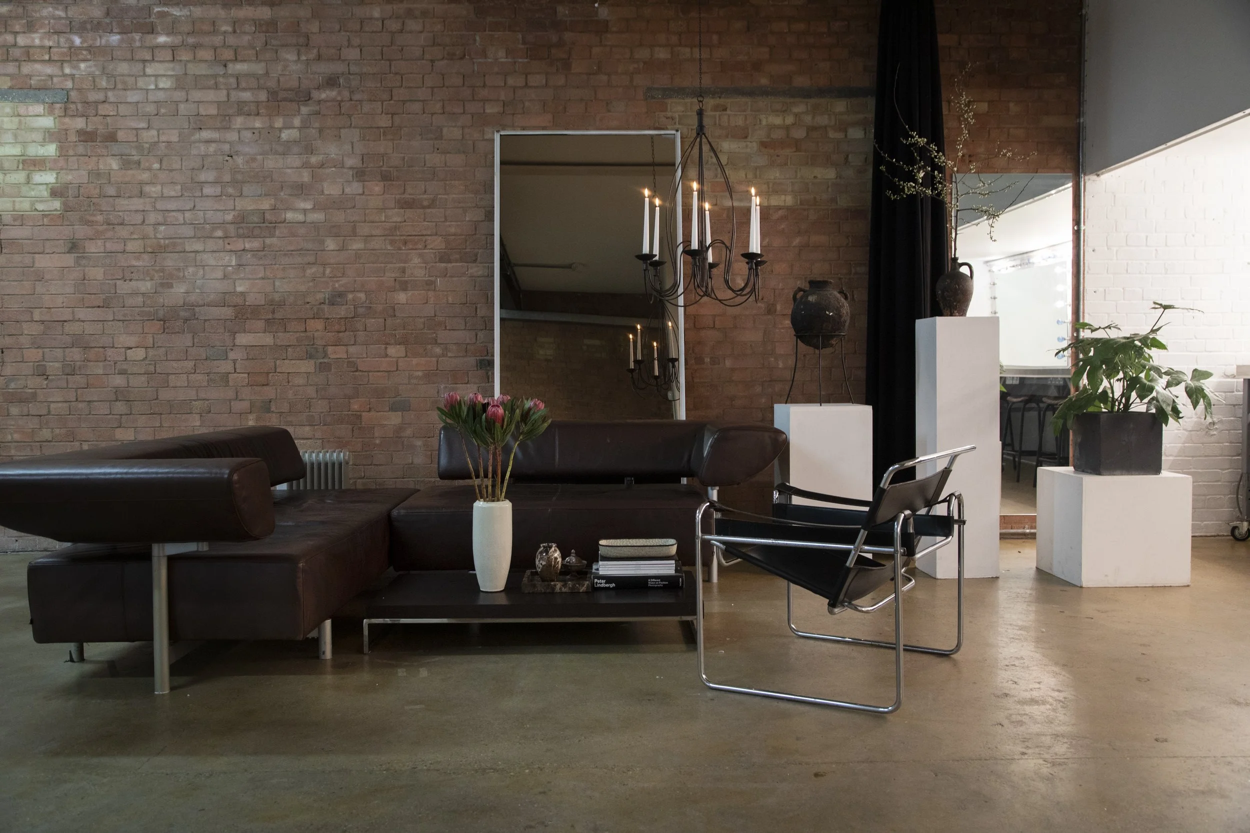 Living room with brick walls, a black leather sectional sofa, a modern black lounge chair, a coffee table with books and a vase of pink tulips, a large mirror, and decorative vases on white pedestals, with a chandelier hanging from the ceiling.