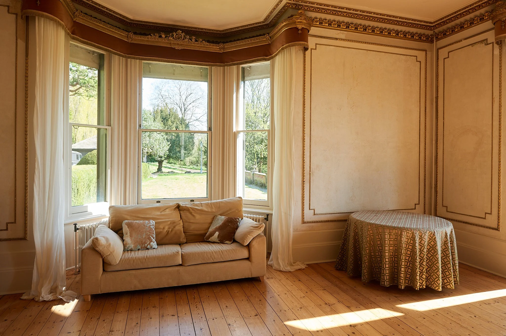 A room with large bay windows, a beige sofa with pillows, and a round table with a patterned tablecloth, overlooking a garden with trees and green grass.