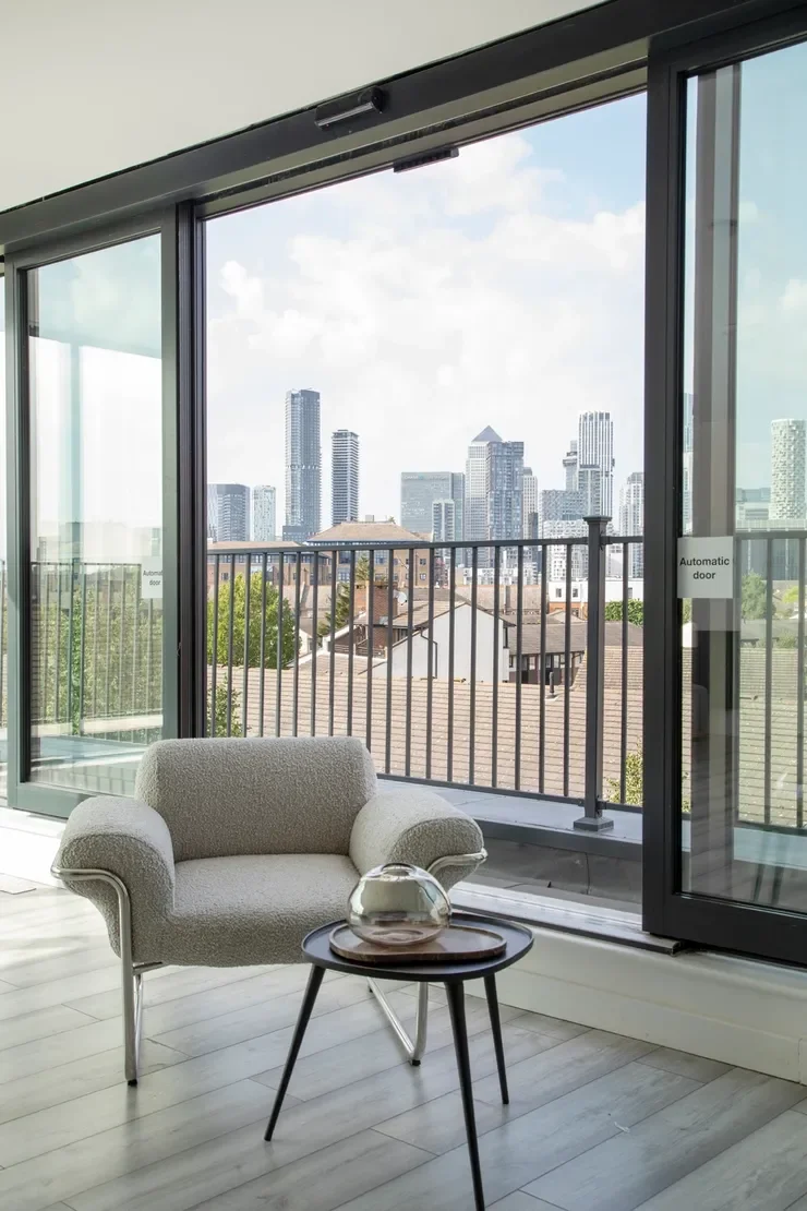 A living room with floor-to-ceiling glass doors opening to a balcony overlooking a city skyline with various tall buildings and a partly cloudy sky.