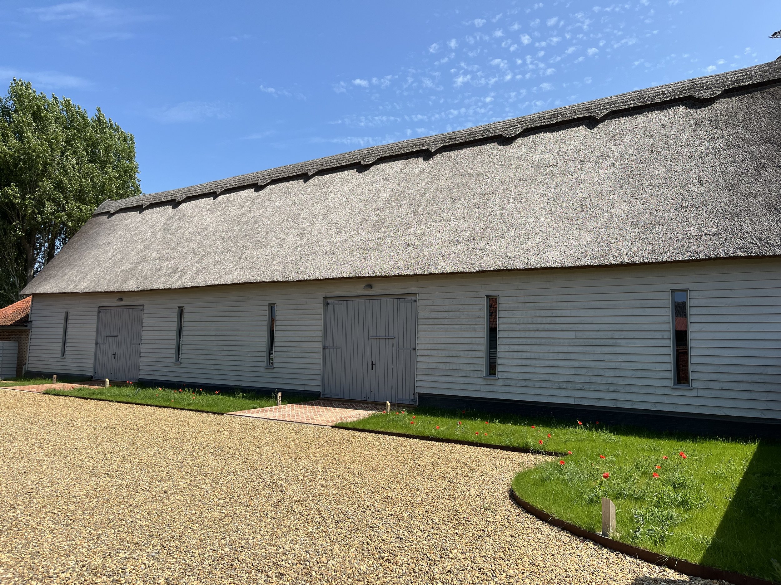 A white wooden building with a thatched roof and narrow windows, surrounded by a gravel pathway and a small grassy area with red flowers, under a blue sky with scattered clouds.