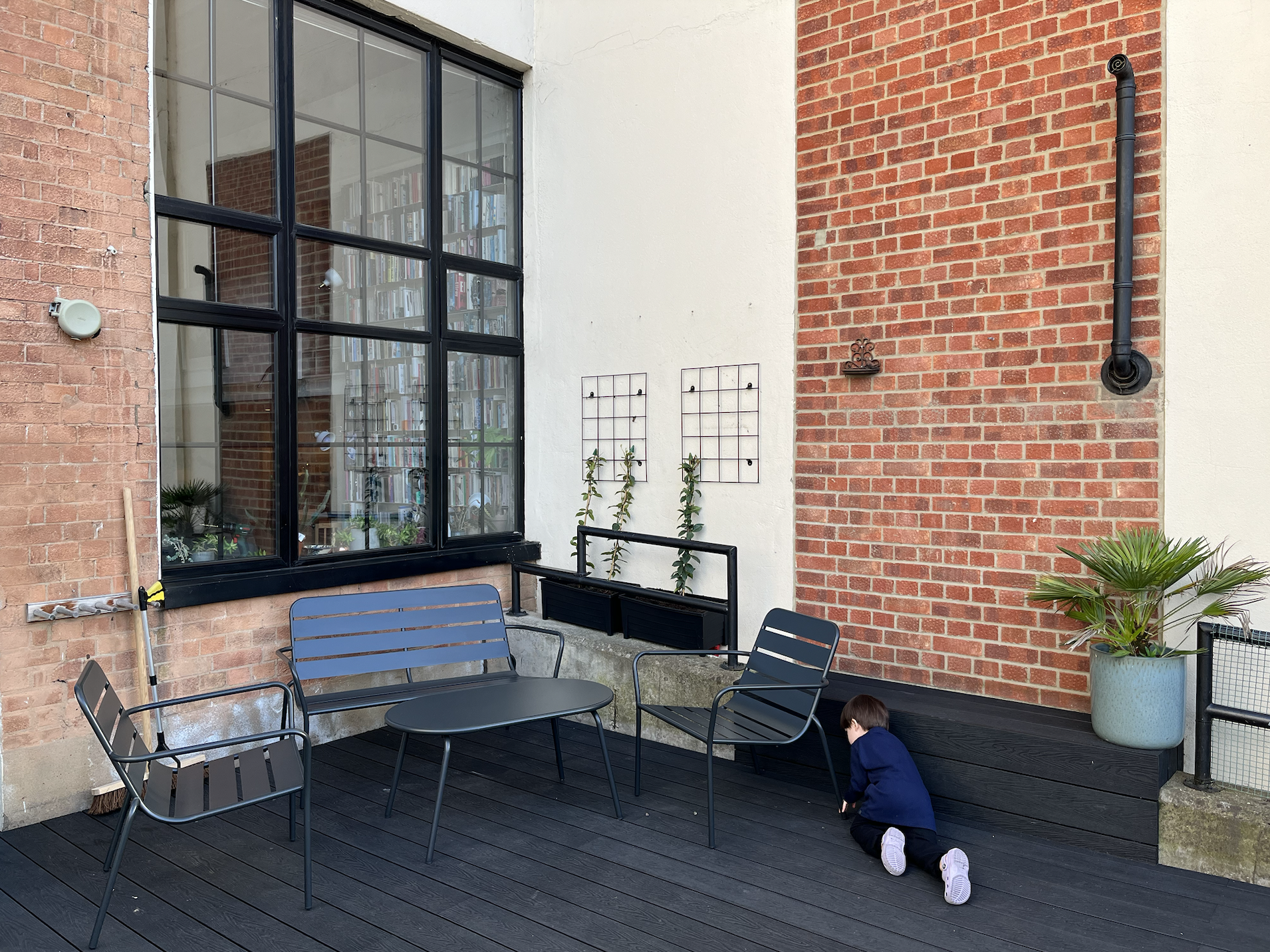 A young boy in a blue jacket crouches down on a dark wooden patio near a large window with black framing, adjacent to a brick wall and white wall, with patio furniture, potted plants, and outdoor wall decor.