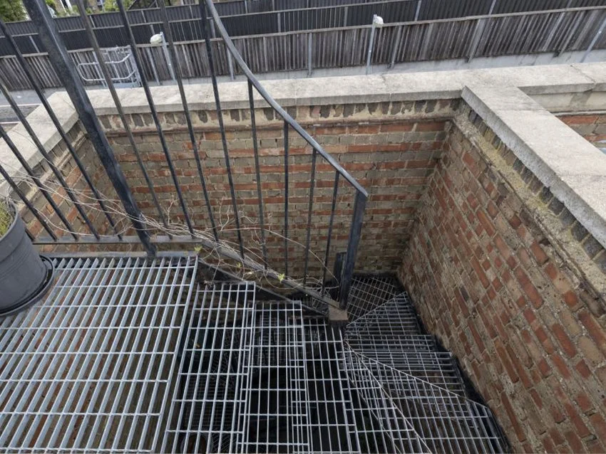 View of an outdoor metal fire escape staircase with a black railing, leading down into a brick-lined basement area, with a potted plant on the metal platform.