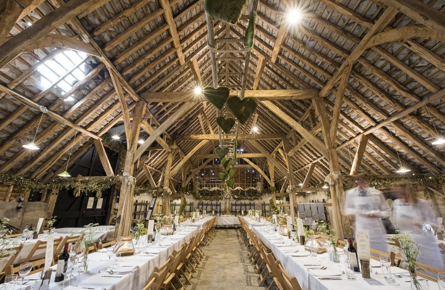 Long rectangular banquet tables set for a wedding reception inside a rustic barn with wooden beams and high vaulted ceiling, decorated with greenery and fairy lights, with some blurred figures of waitstaff serving.