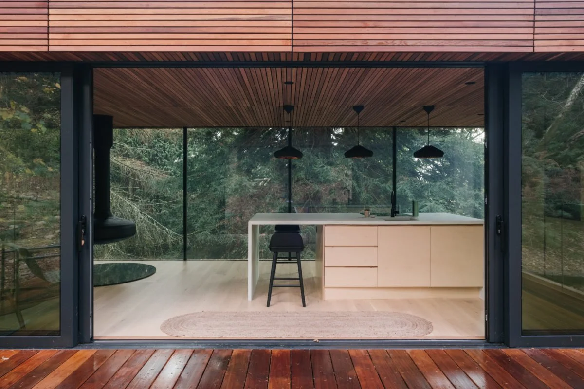 Modern kitchen with a white island and black pendant lights, large glass sliding doors, and a wooded outdoor view.