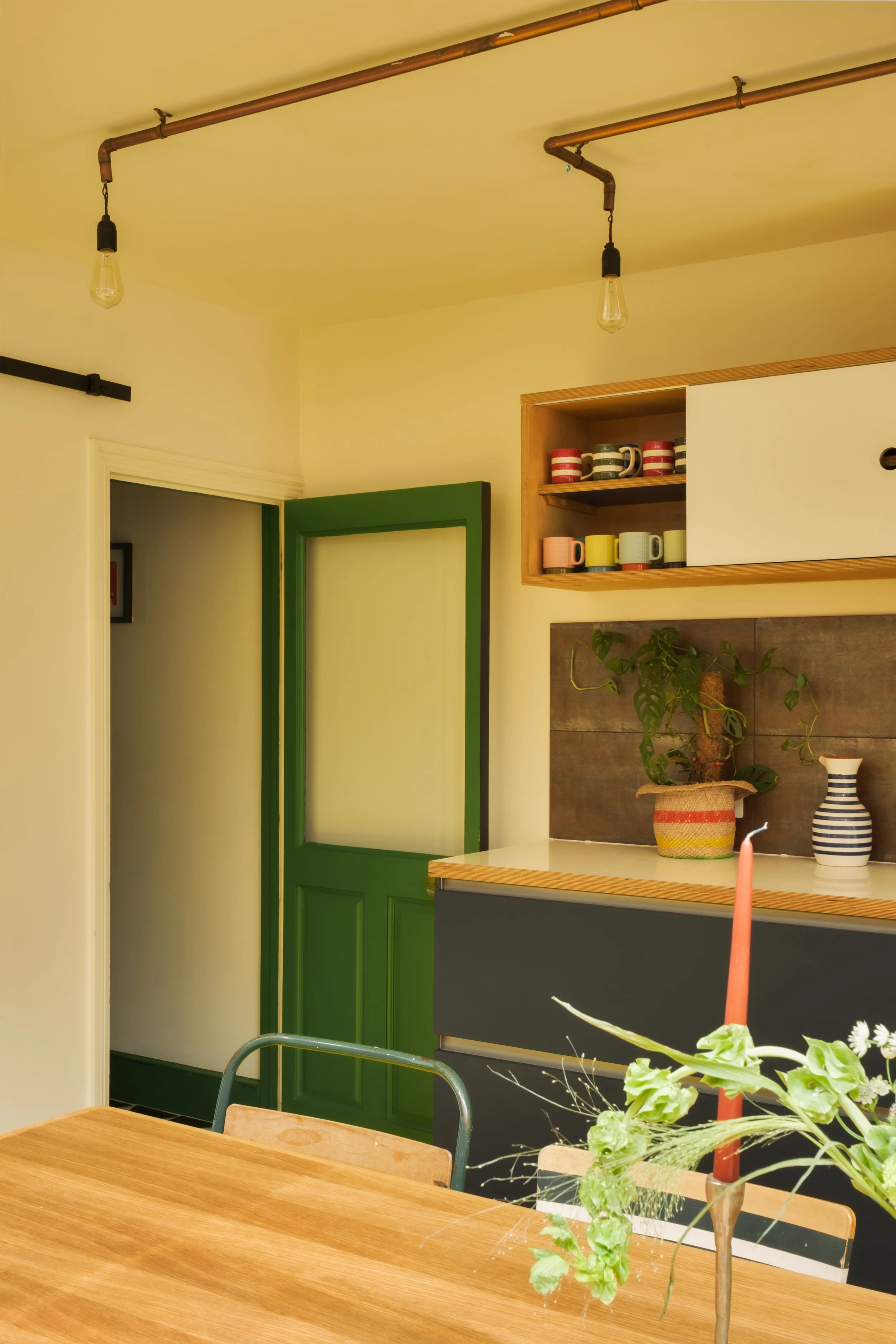 Kitchen with a wooden table, decorative plants, colorful cups on shelf, green door, and exposed light bulbs on ceiling.