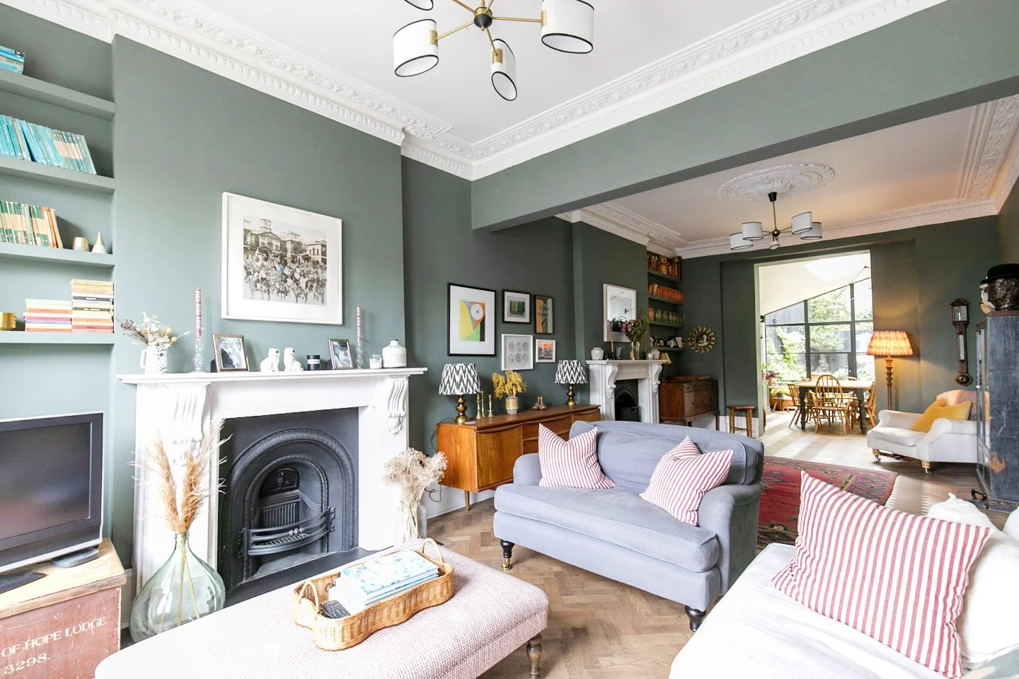 Interior of a living room with green walls, white fireplace, wooden furniture, a gray sofa with striped pillows, a white armchair, and a dining area in the background, decorated with artwork and lamps.