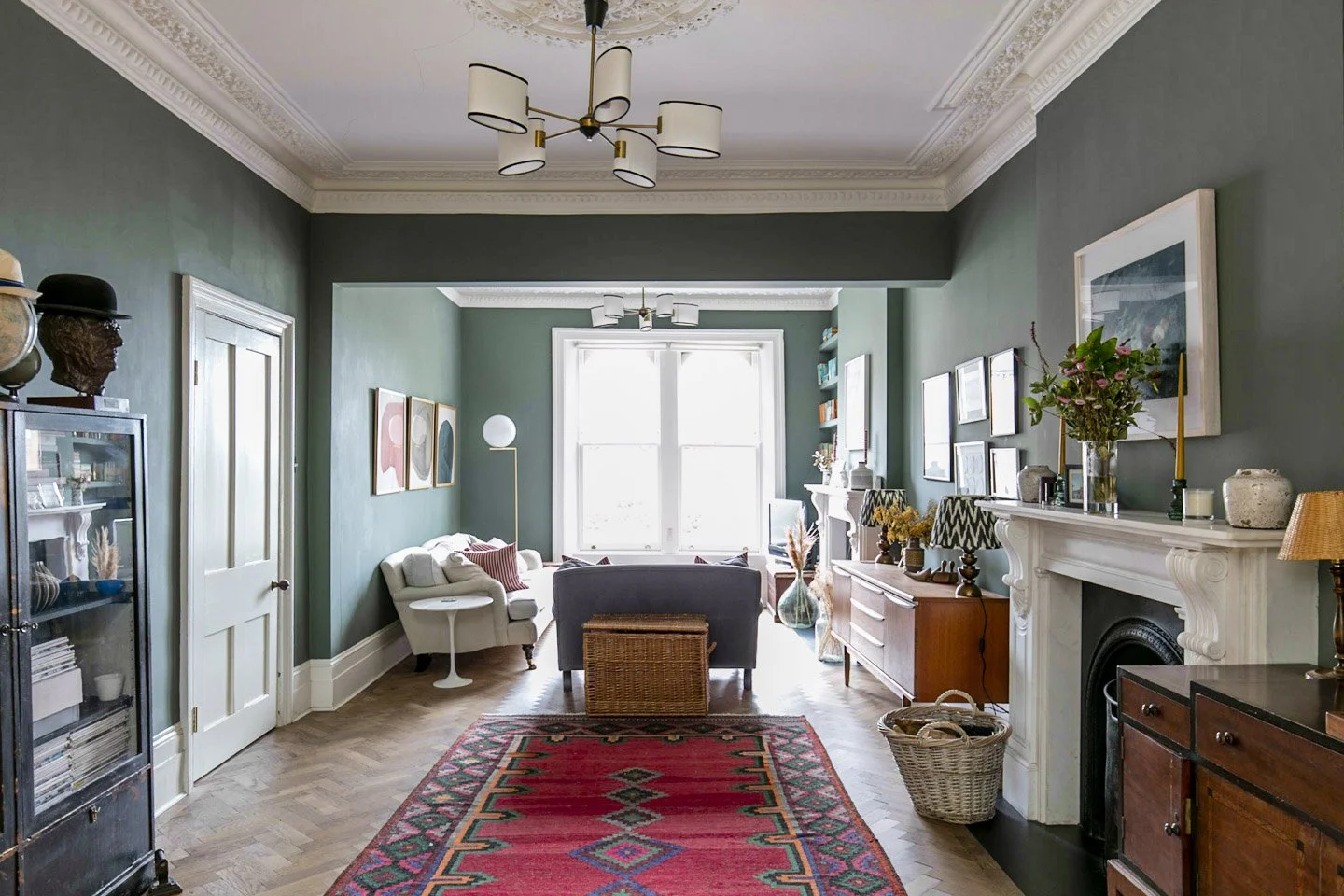 Living room with green walls, white molding, large window, mix of vintage and modern furniture, colorful artwork, patterned area rug, and decorative items.