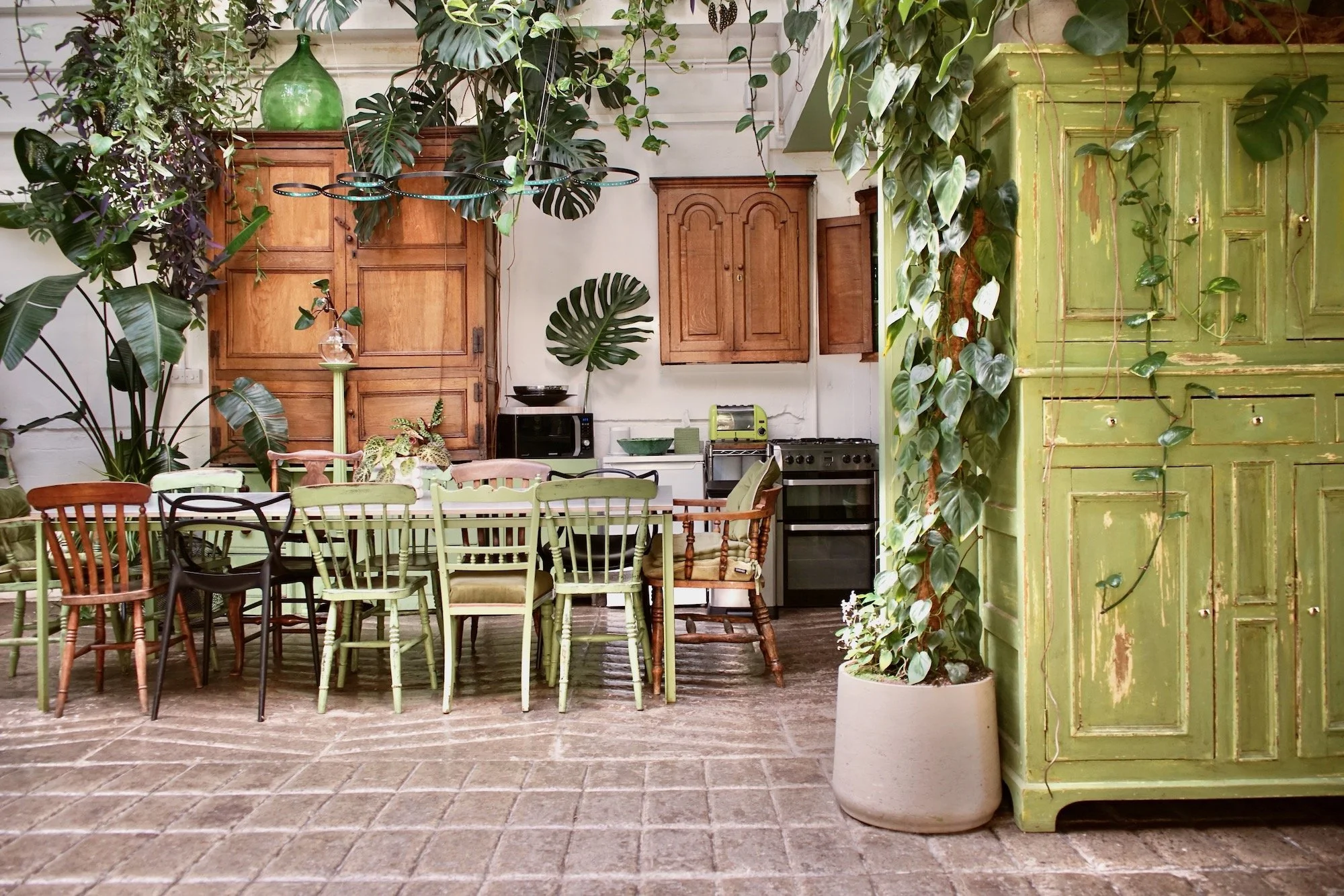 A cozy kitchen and dining area with green and wooden furniture, surrounded by green plants and a brick floor.