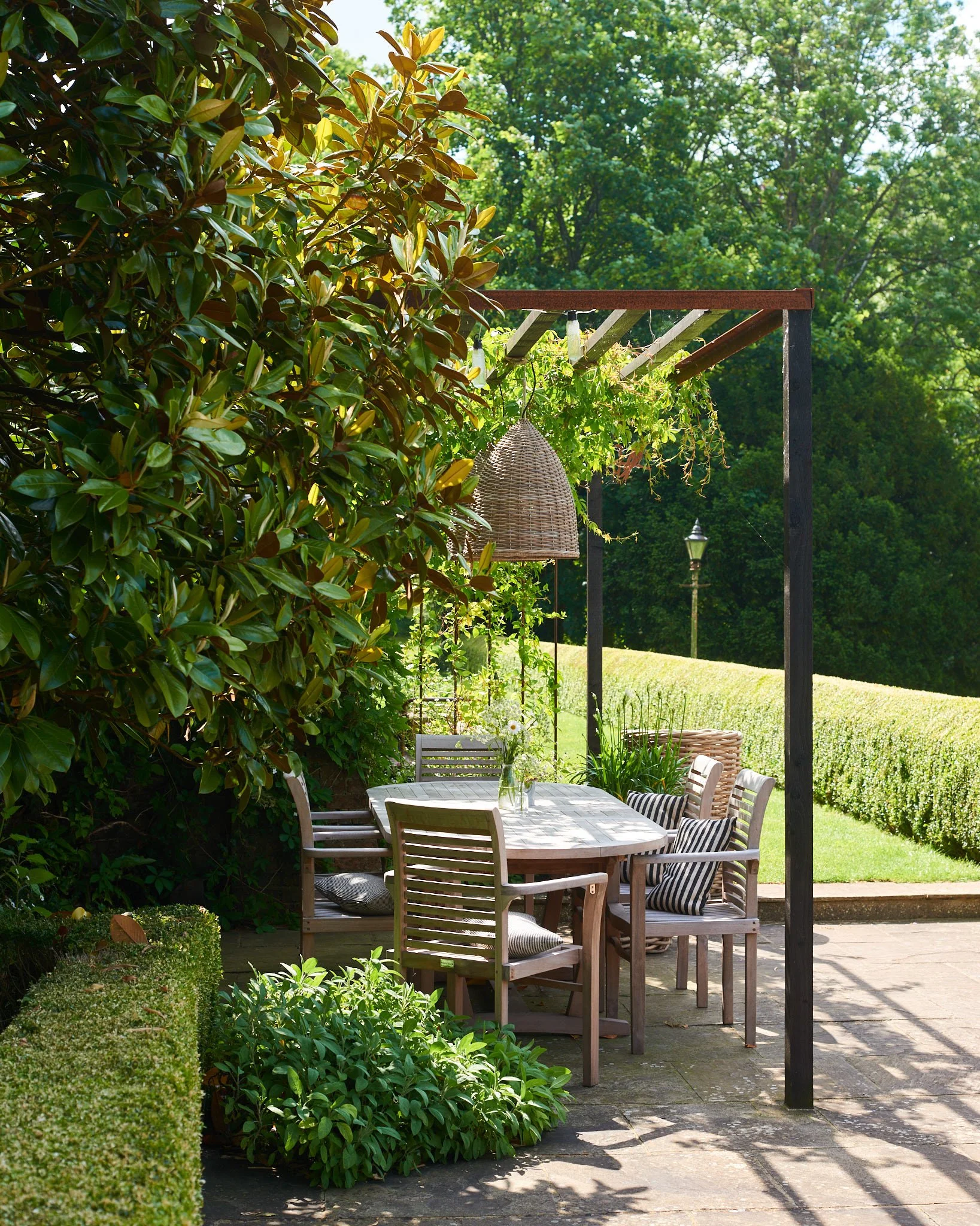 Outdoor patio with a wooden dining table, six chairs with cushions, under a wooden pergola with hanging wicker light fixture, surrounded by lush greenery and hedges, with a garden and trees in the background.