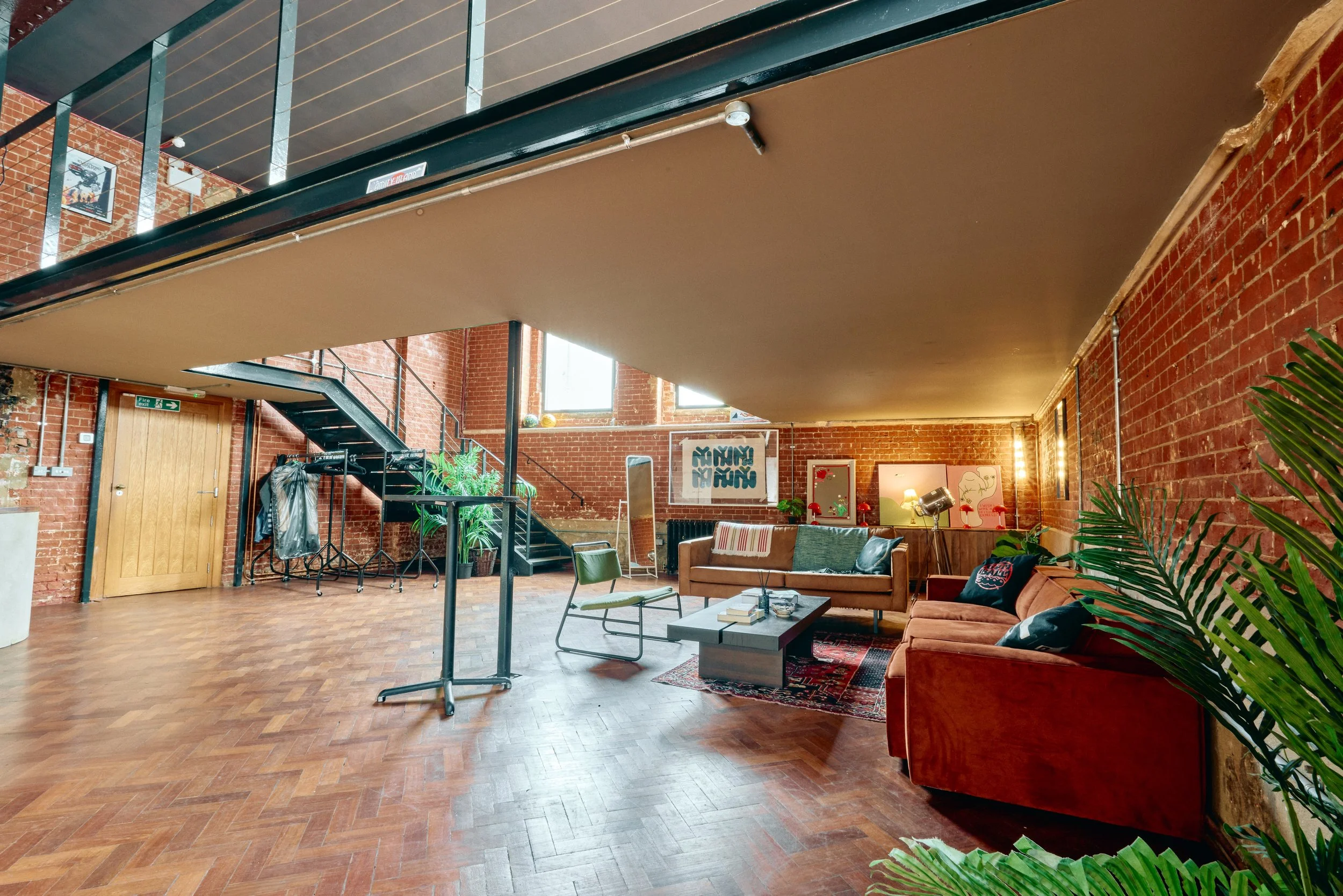 A spacious living room with exposed red brick walls, wooden flooring, and large windows. It features a brown couch with pillows, a coffee table, a green chair, and several decorative plants. There are modern art pieces on the wall and a staircase lea