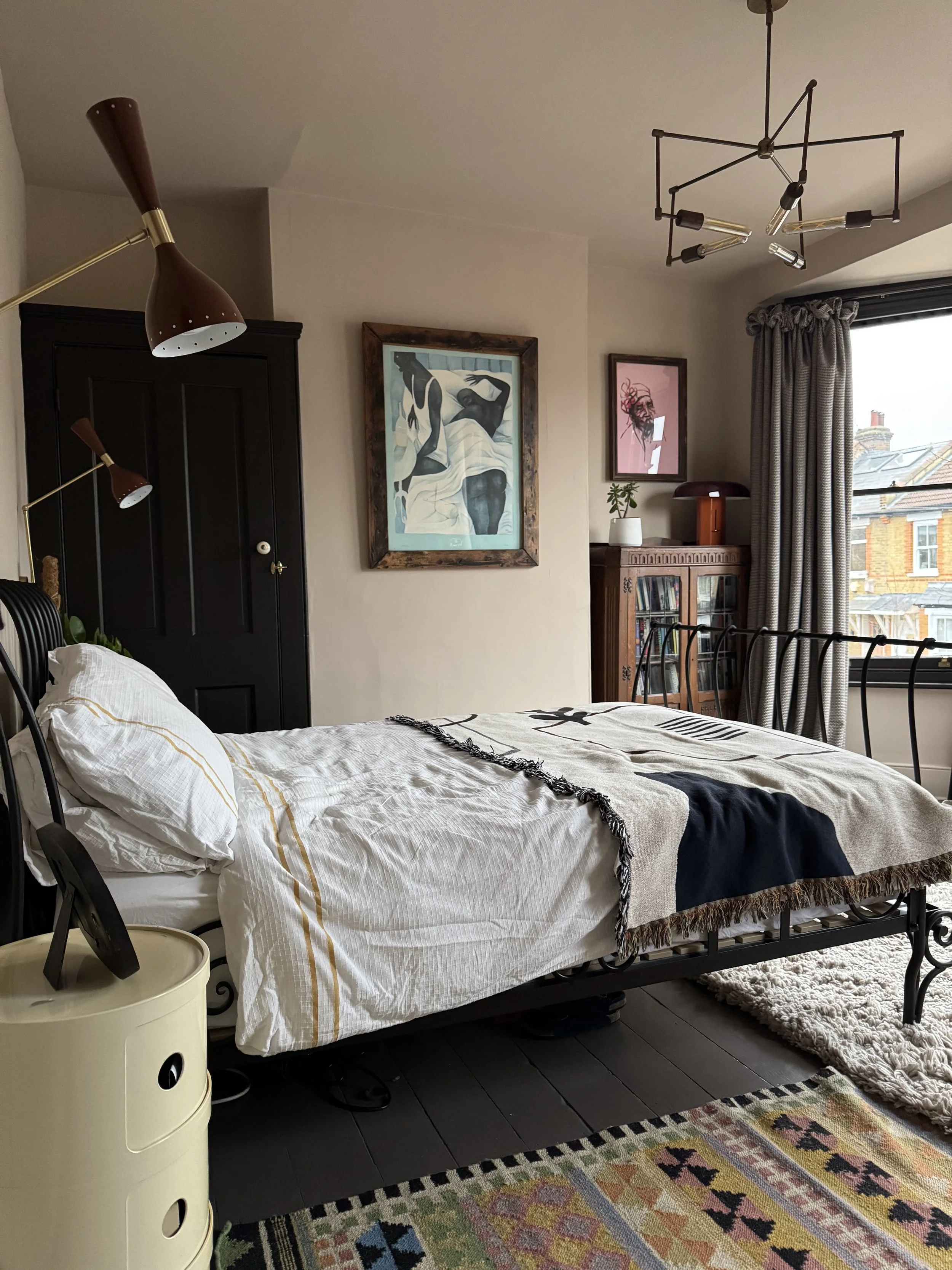 A bedroom with a black bed frame, white and beige bedding, and a patterned blanket. There is a bedside lamp and a cream-colored bedside table with a black clock. The wall has two framed art pieces, one abstract and one portrait, and a wooden cabinet 