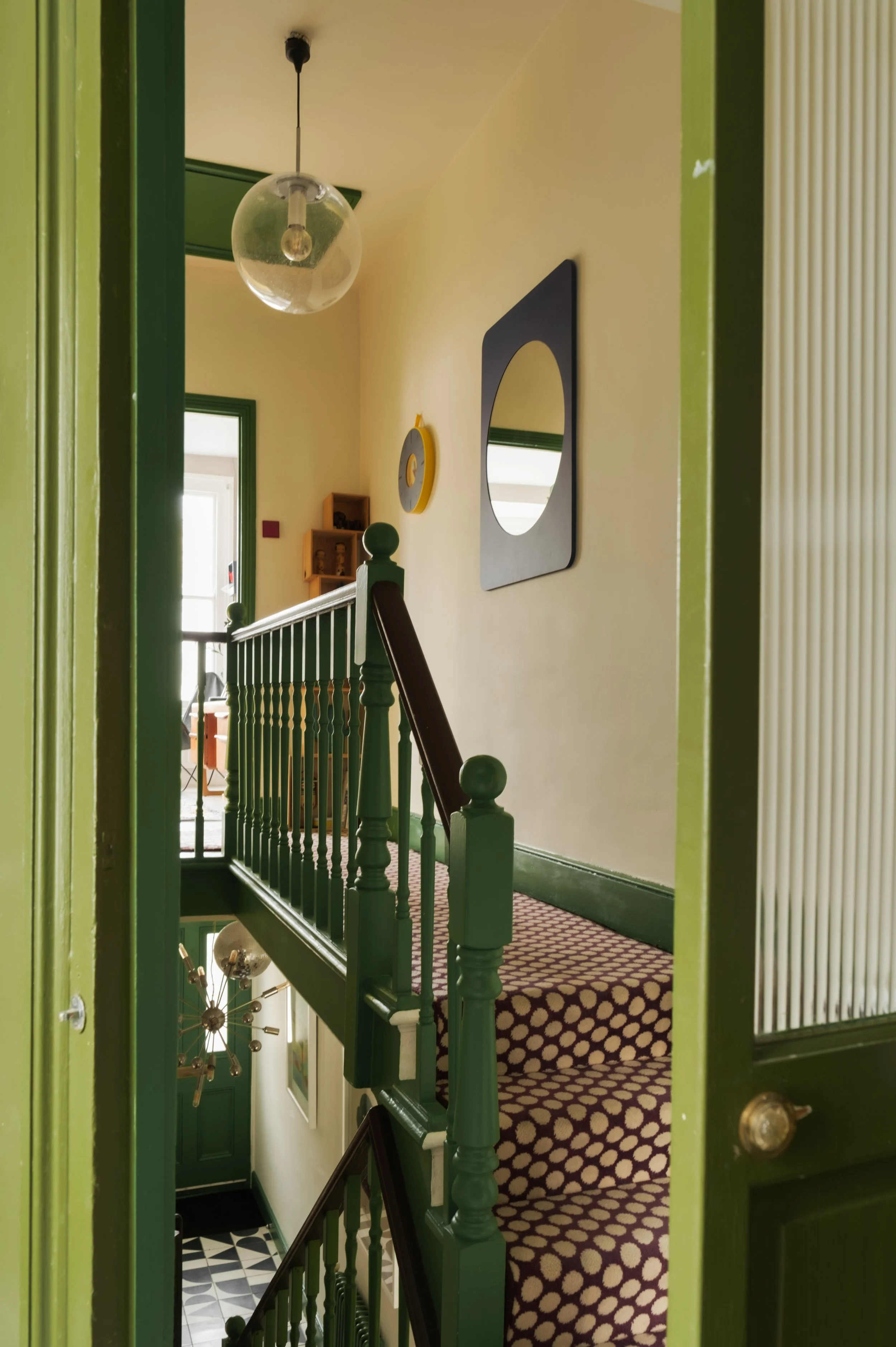 Interior view of a staircase landing with patterned carpet, green wooden railings, and yellow walls with modern wall art and a hanging glass globe light fixture.