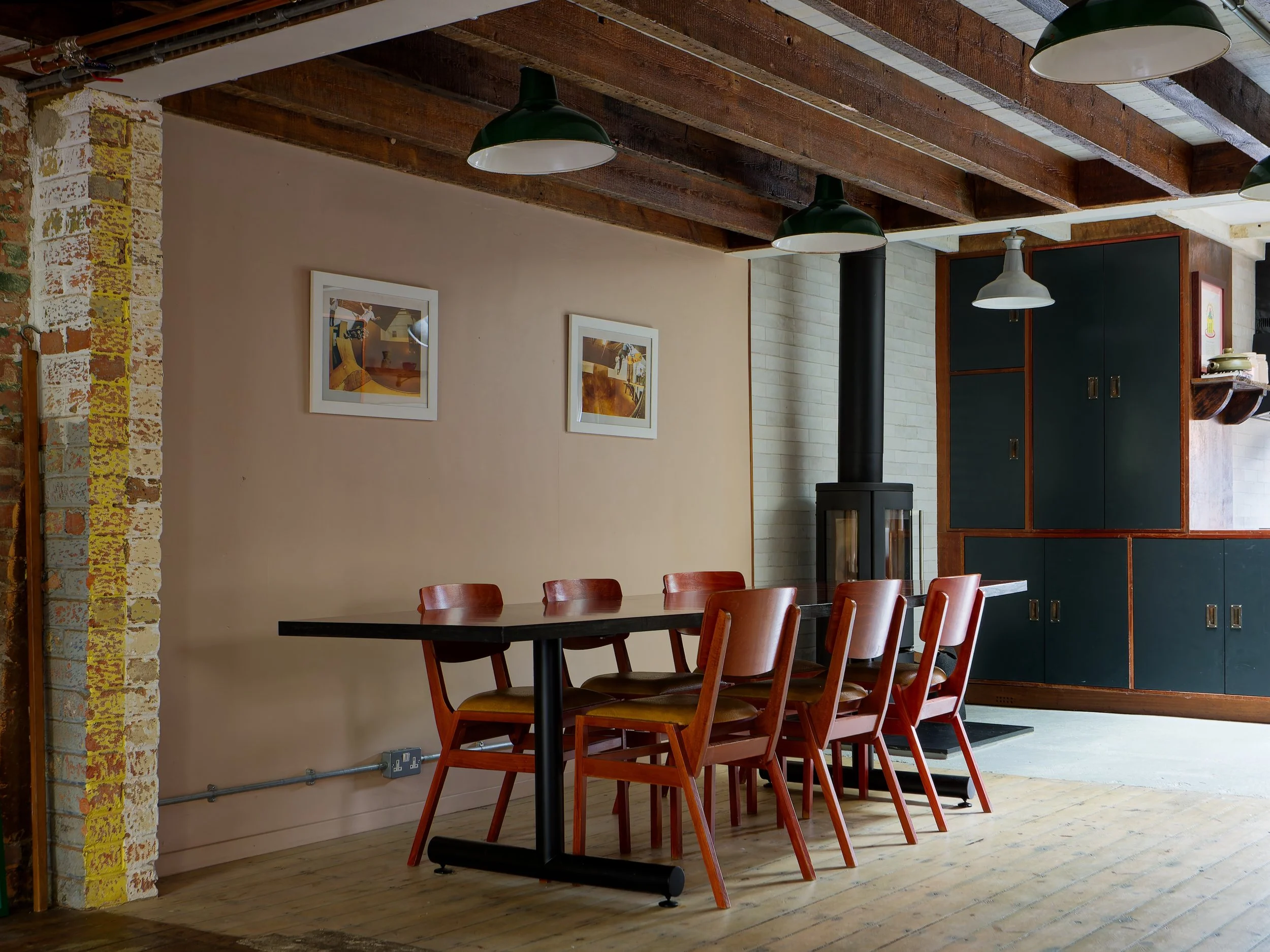 Interior of a cozy dining area with a black rectangular table surrounded by six wooden chairs, a wood stove in the background, and a dark green cabinet with wooden framing along the wall. The ceiling has exposed wooden beams and three large black and