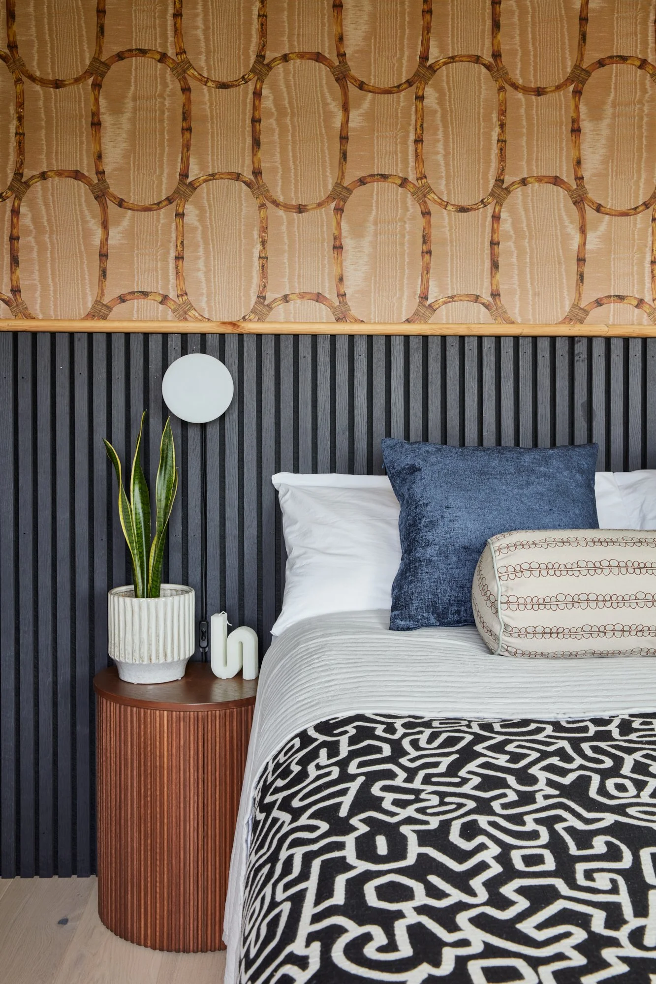 A bedroom with a black paneled headboard, white bedding, a dark blue pillow, a cylindrical decorative pillow, a wooden side table with a potted snake plant, a white s-shaped decor piece, and a round wall sconce light. The wall above the headboard fea
