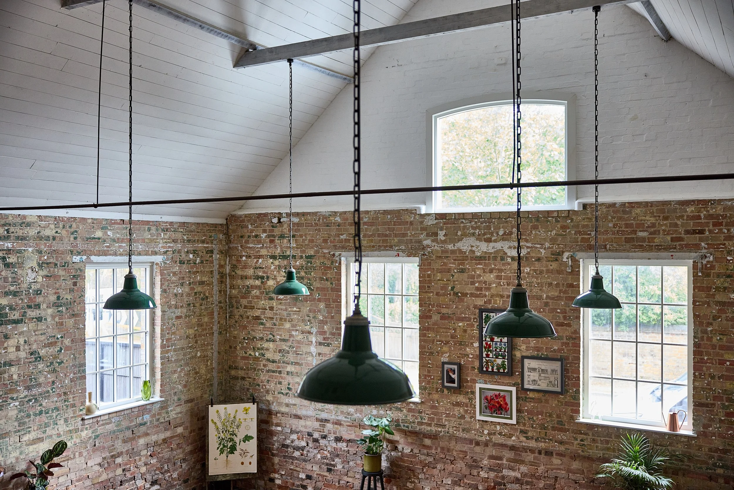 Interior of a brick-walled room with large windows, hanging green industrial-style pendant lights, and framed artwork on the wall. Plants are on the windowsills and floor.