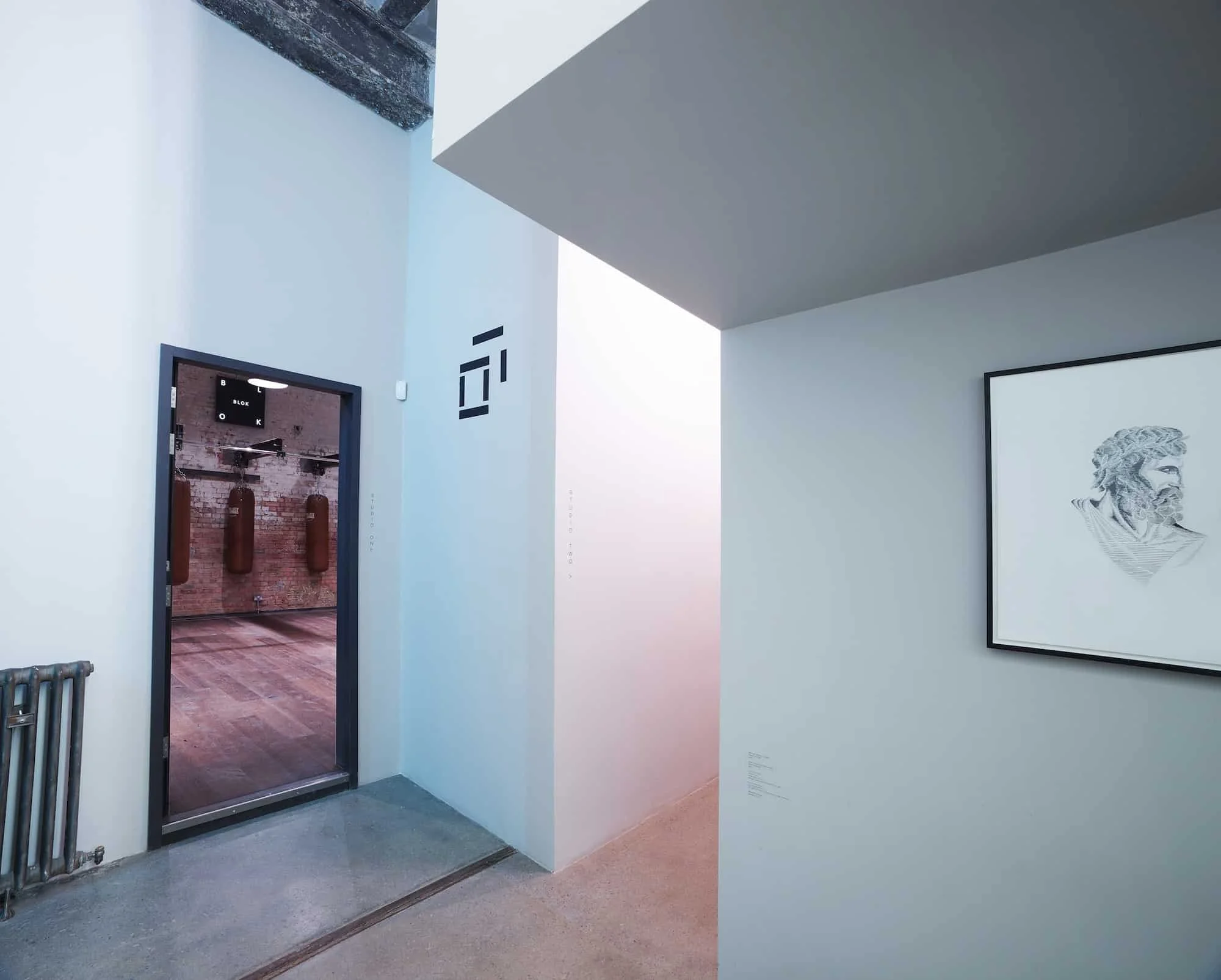 Art gallery interior with framed monochrome portrait on right wall, doorway leading to room with punching bags and exposed brick wall, minimalist decor, and soft lighting.