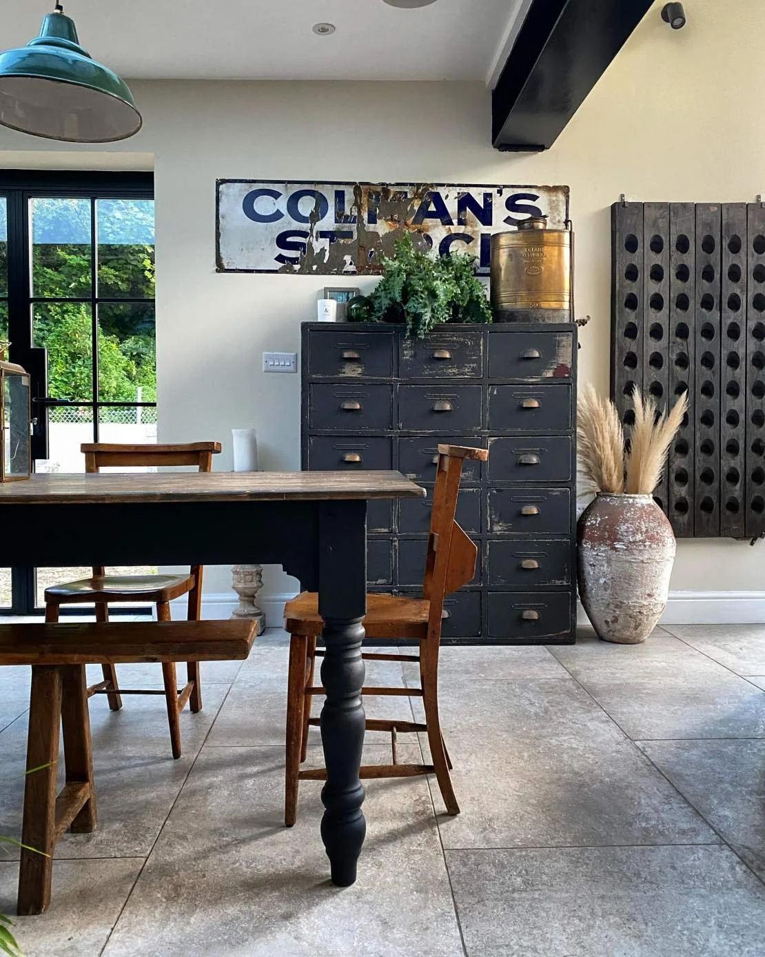 A rustic dining room with a large wooden table and chairs, a black metal cabinet with drawers, a vintage sign reading 'Colman's', greenery, decorative vase with pampas grass, and a window showing a green outdoor scene.