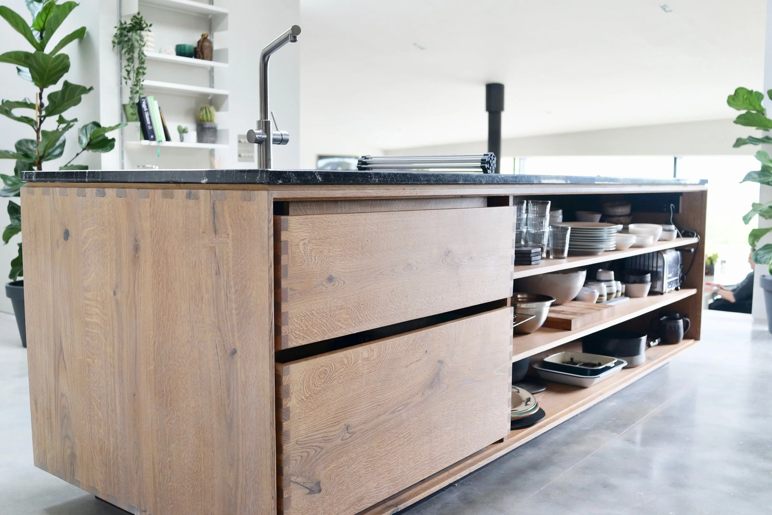 Modern kitchen island with open shelving and a black countertop, surrounded by green plants and decorated with dishes and kitchenware.