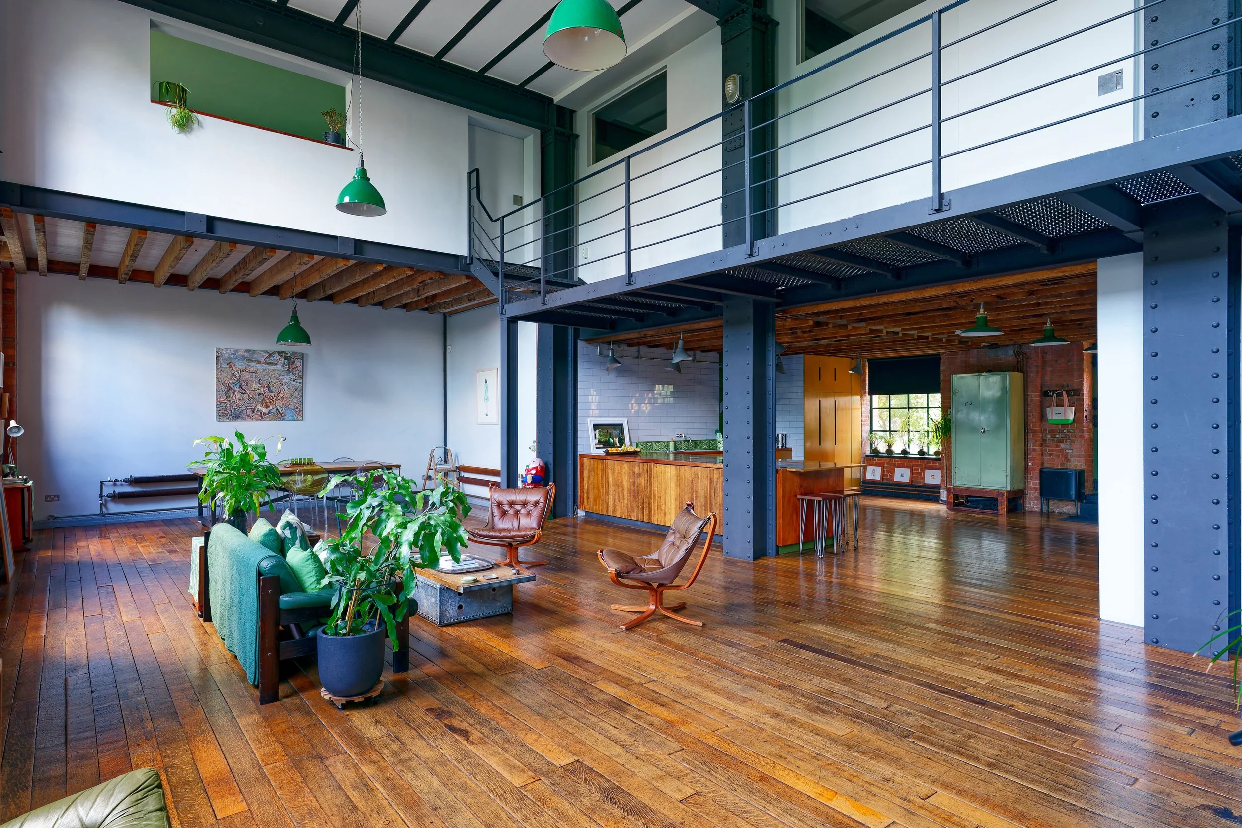 Open indoor living space with high ceiling and extensive wooden flooring, featuring a mix of modern and industrial design elements including green pendant lights, potted plants, leather chairs, and a wooden bar area with stools.