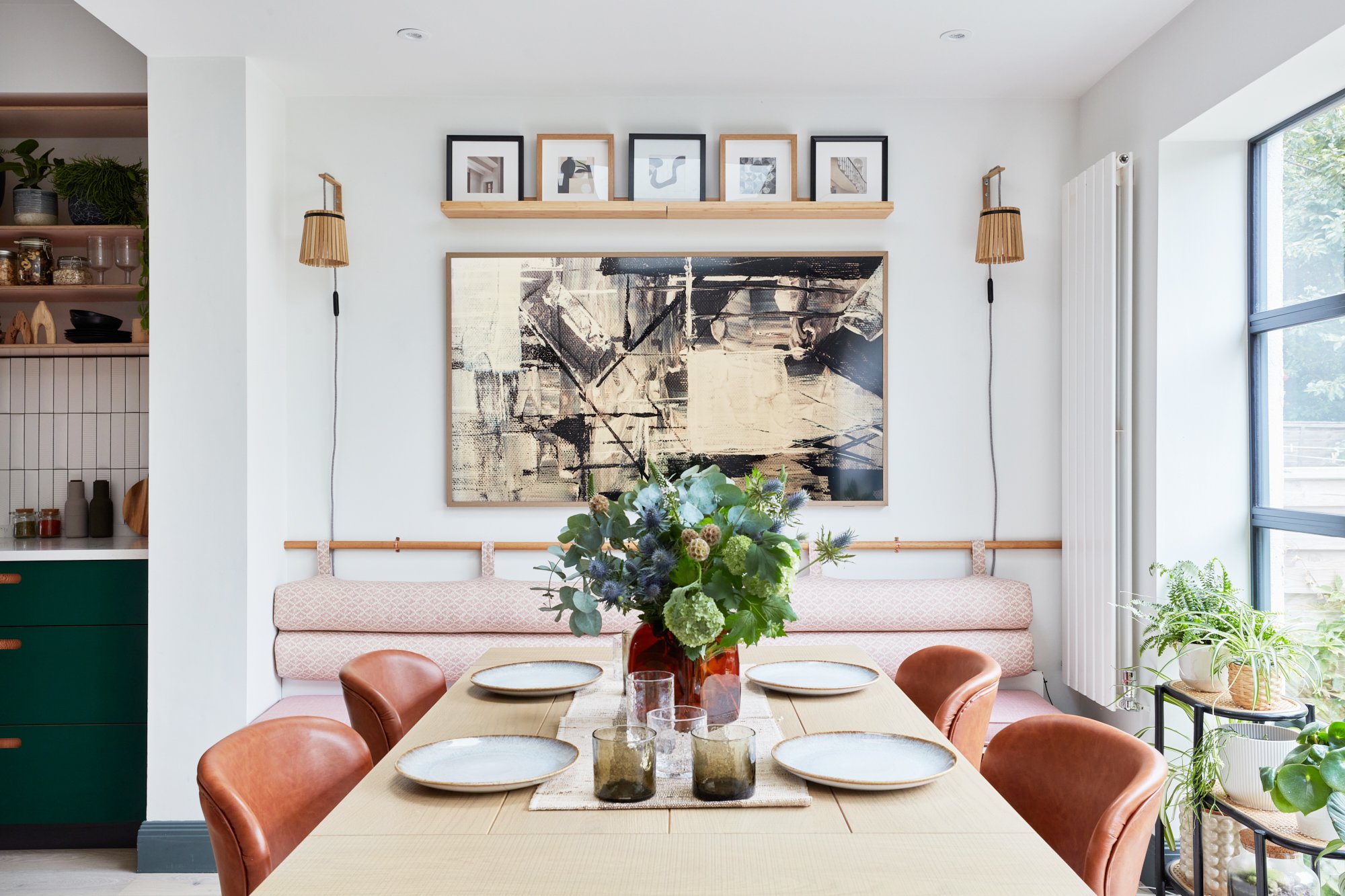 A dining area with a rectangular table set with six plates and four glasses, centered with a floral arrangement in a red vase, surrounded by six brown leather chairs. Behind the table is a built-in bench with padded pink upholstery and a large black-