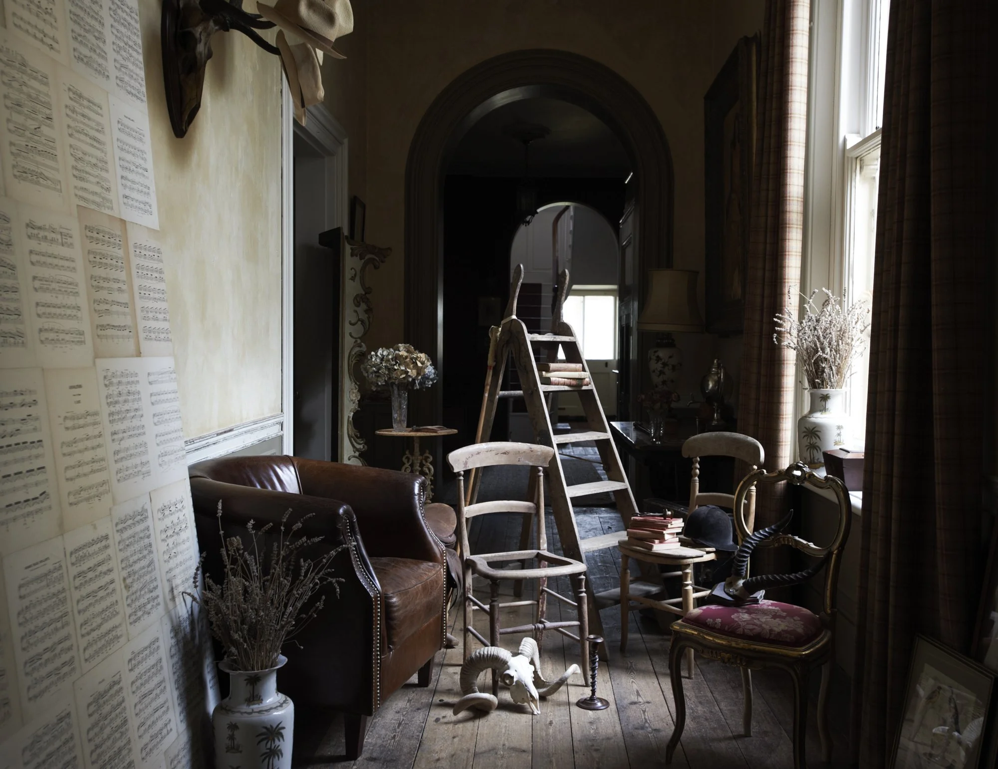 An interior room with vintage furniture, a ladder, books, and decorative items, lit by natural light from large windows with curtains.