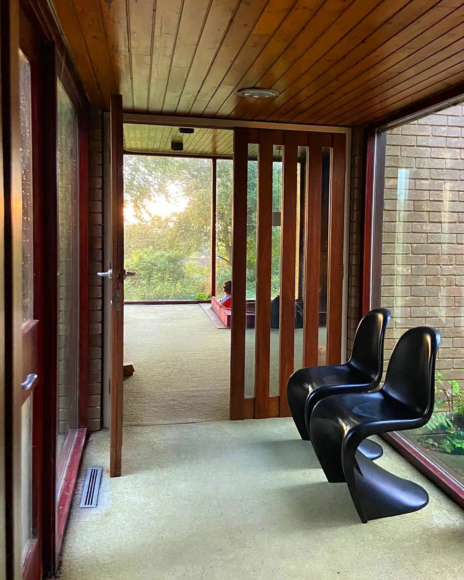 A sunlit enclosed porch area featuring two black modern chairs near large windows, with a woman sitting on a pink bench outside in the distance, surrounded by greenery.