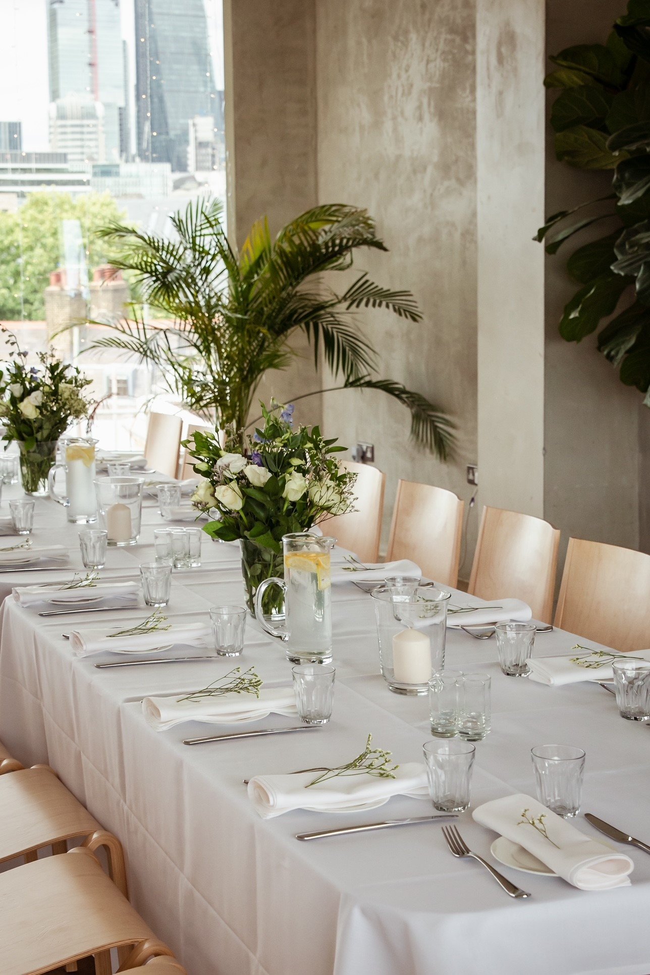 Elegant dining table set with white tablecloth, glassware, silverware, napkins, and floral centerpieces, near a large window with city skyline view.