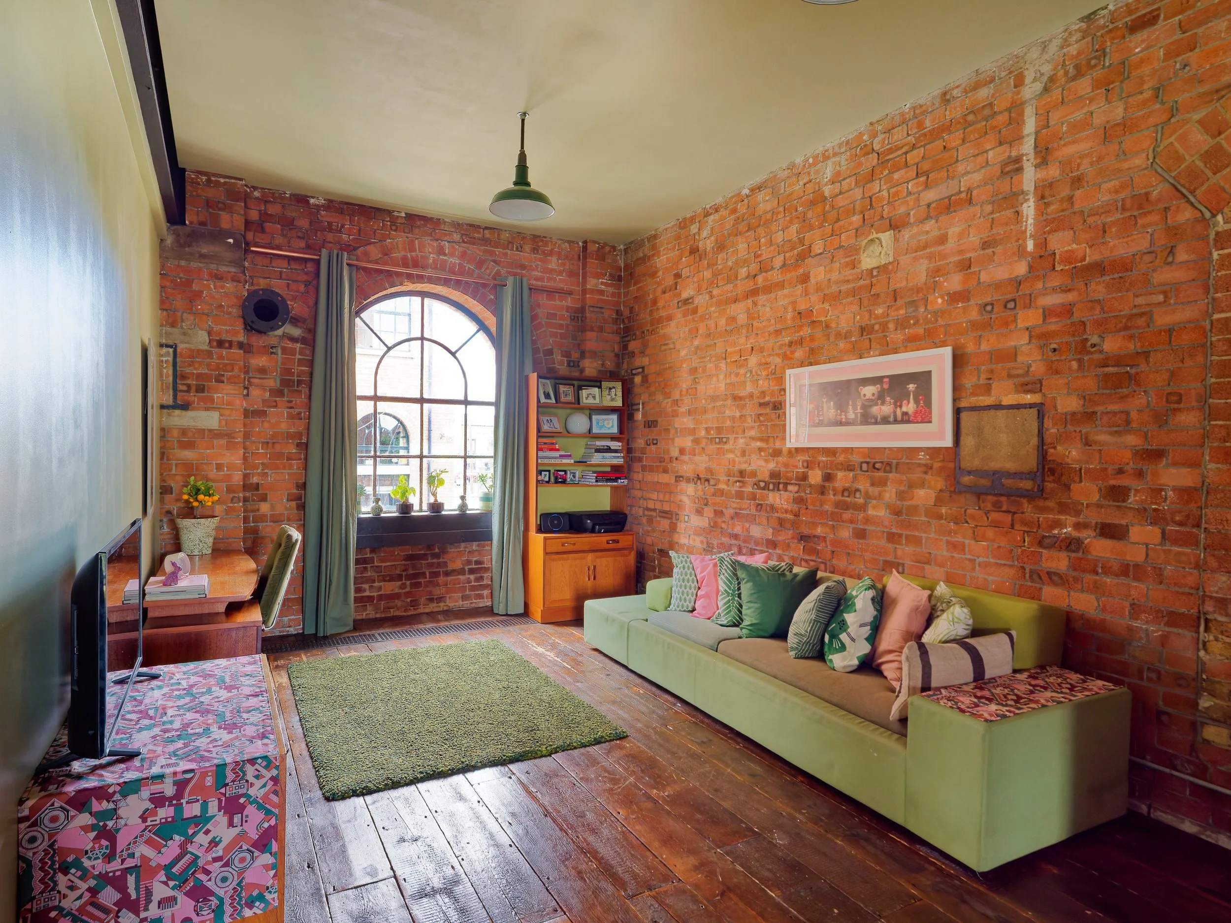 Living room with exposed brick wall, green sofa with multiple pillows, wooden floor, large arched window with curtains, bookshelf, and small desk.