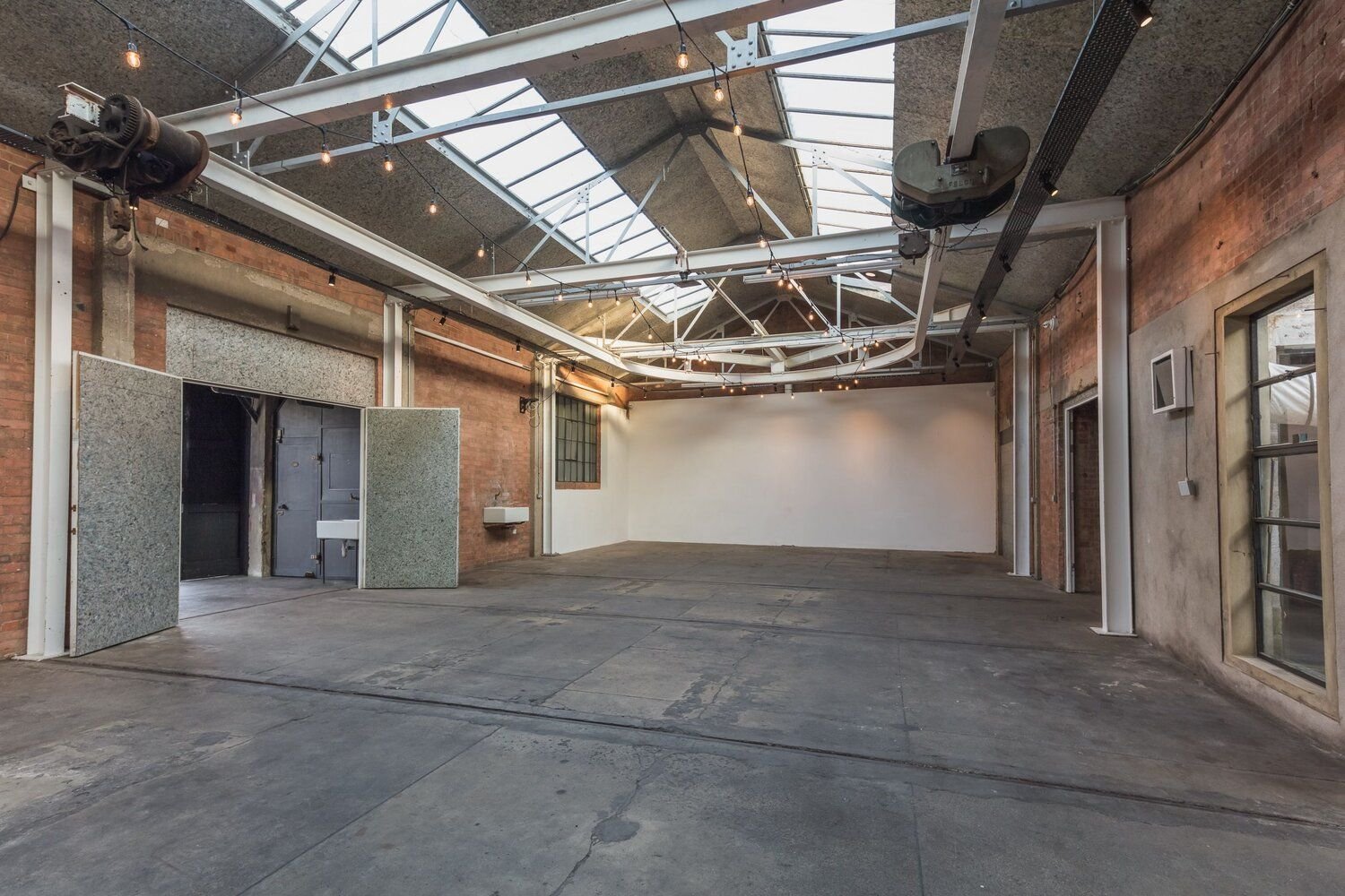 Empty industrial-style event space with exposed brick walls, concrete floor, large skylight ceiling, string lights, and a white wall at the back.