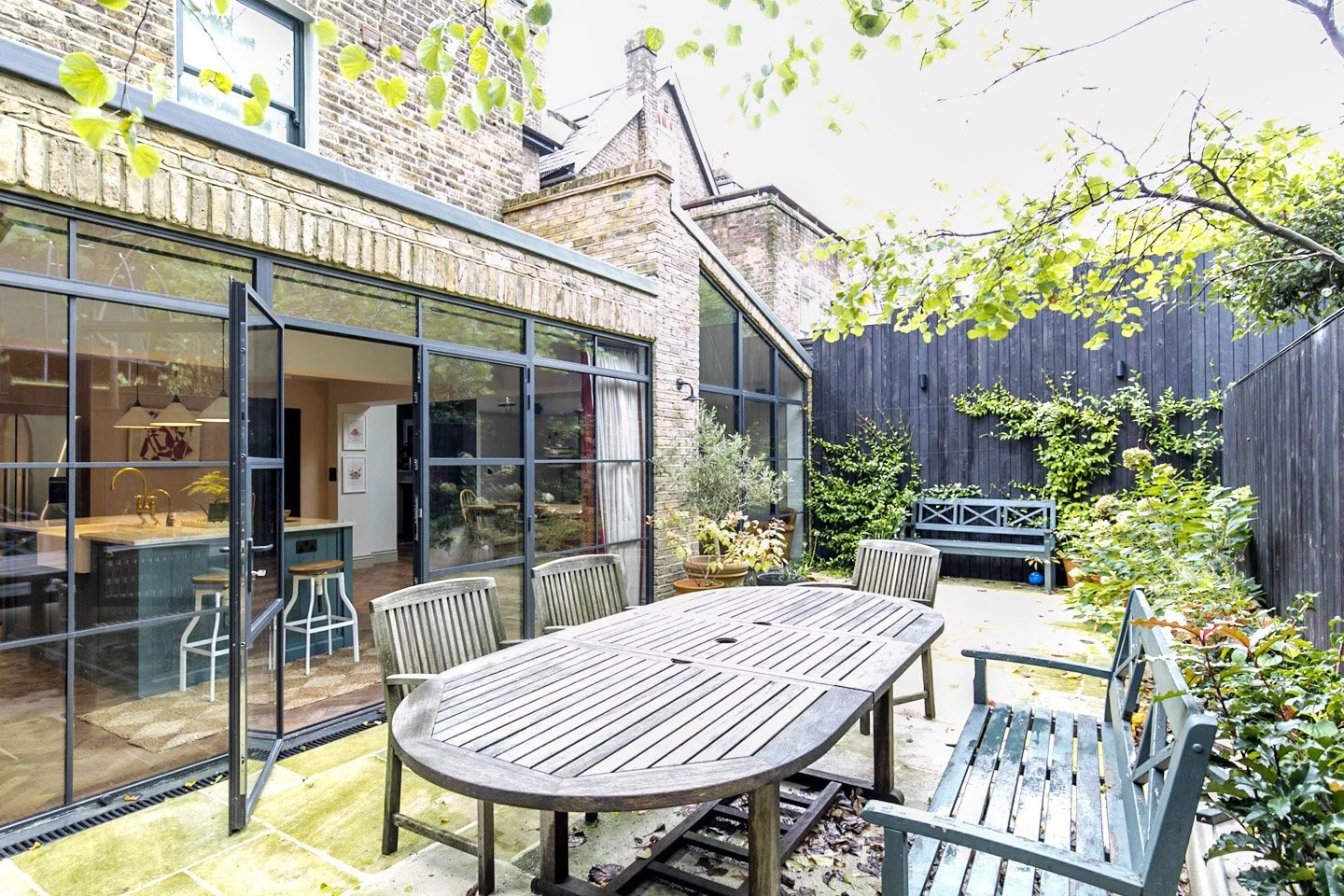 Outdoor patio with a large wooden table, chairs, and benches, enclosed by dark wooden fencing, with potted plants and greenery, and a house with large glass windows revealing the interior kitchen.