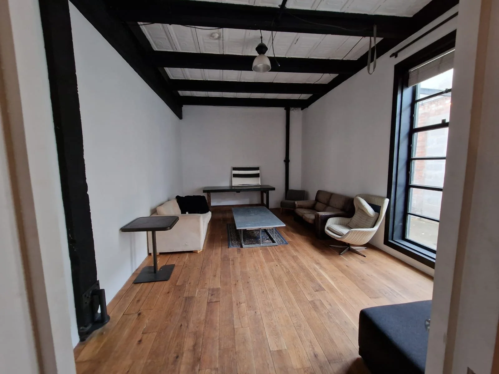 A minimalist living room with white walls, wooden floor, and a high ceiling with black exposed beams. There's a small beige sofa, a dark sofa, an armchair with a swivel base, and a small coffee table. Window on the right side letting in natural light