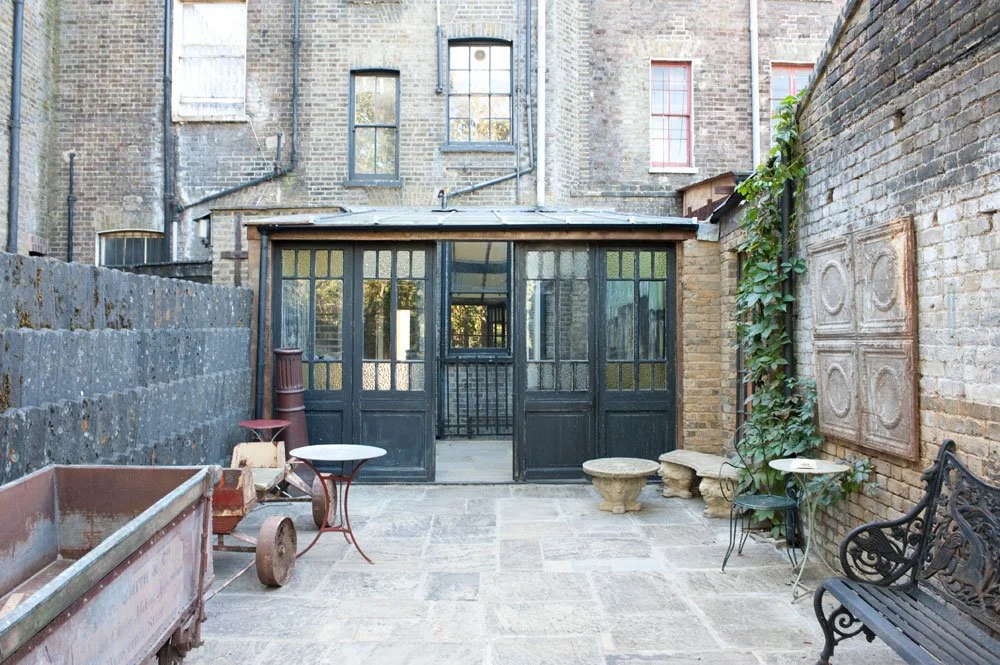 Urban backyard with stone pavement, brick walls, a black glass enclosed structure, outdoor furniture including stone benches and metal tables, and climbing plants on the wall.