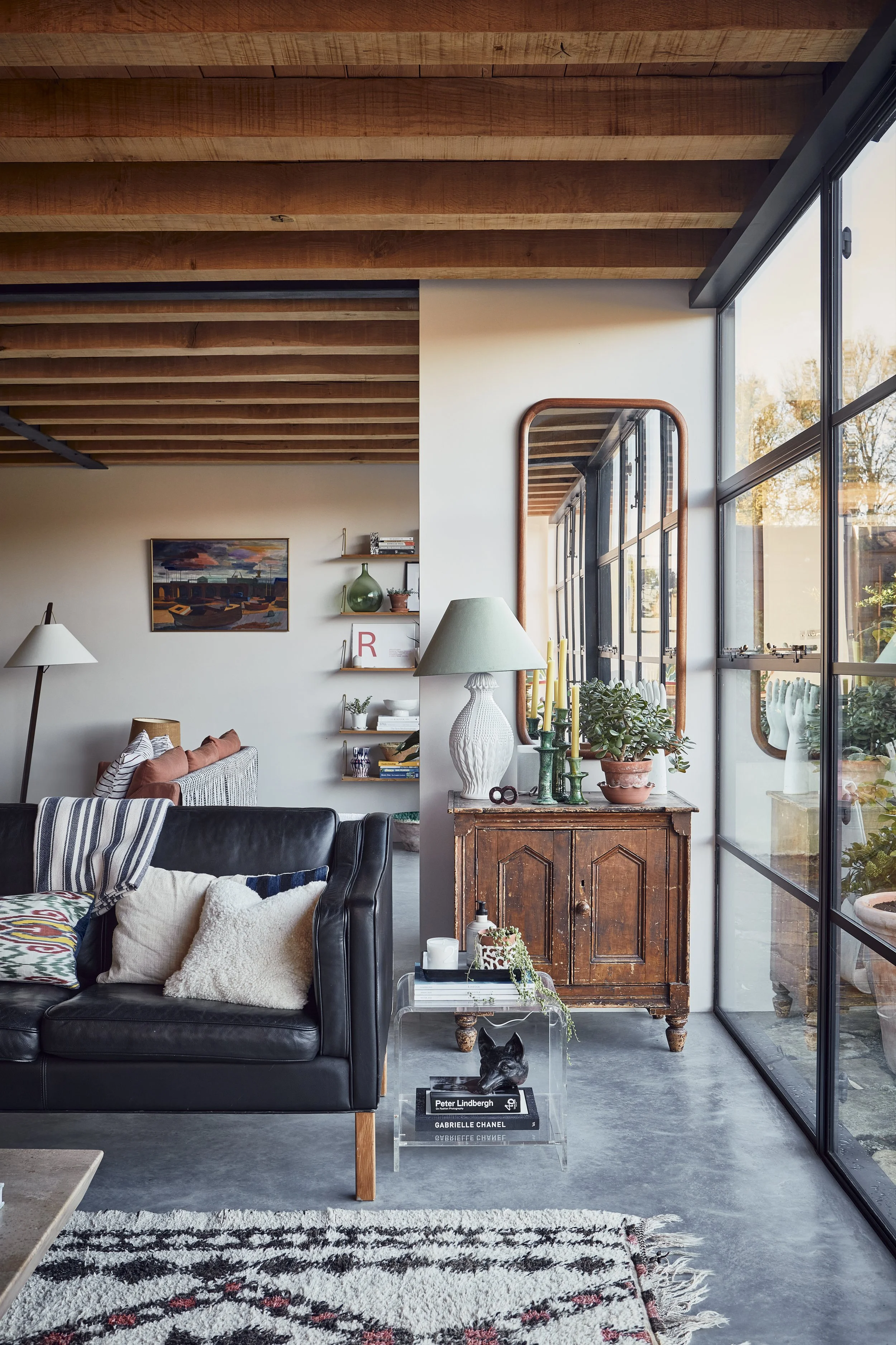 A modern living room featuring a black leather sofa with patterned and plain pillows, a glass side table with books and a dog sculpture, a vintage wooden cabinet with decorative plants and candles, large floor-to-ceiling glass windows, and wooden cei