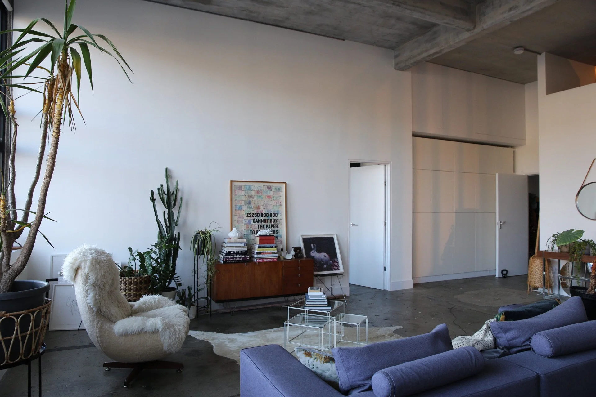 Modern living room with white walls, concrete floor, and ceiling featuring exposed concrete beams. Contains a large potted plant, a fluffy white armchair, a purple sofa, a wooden sideboard with books and art, and various framed pictures leaning again