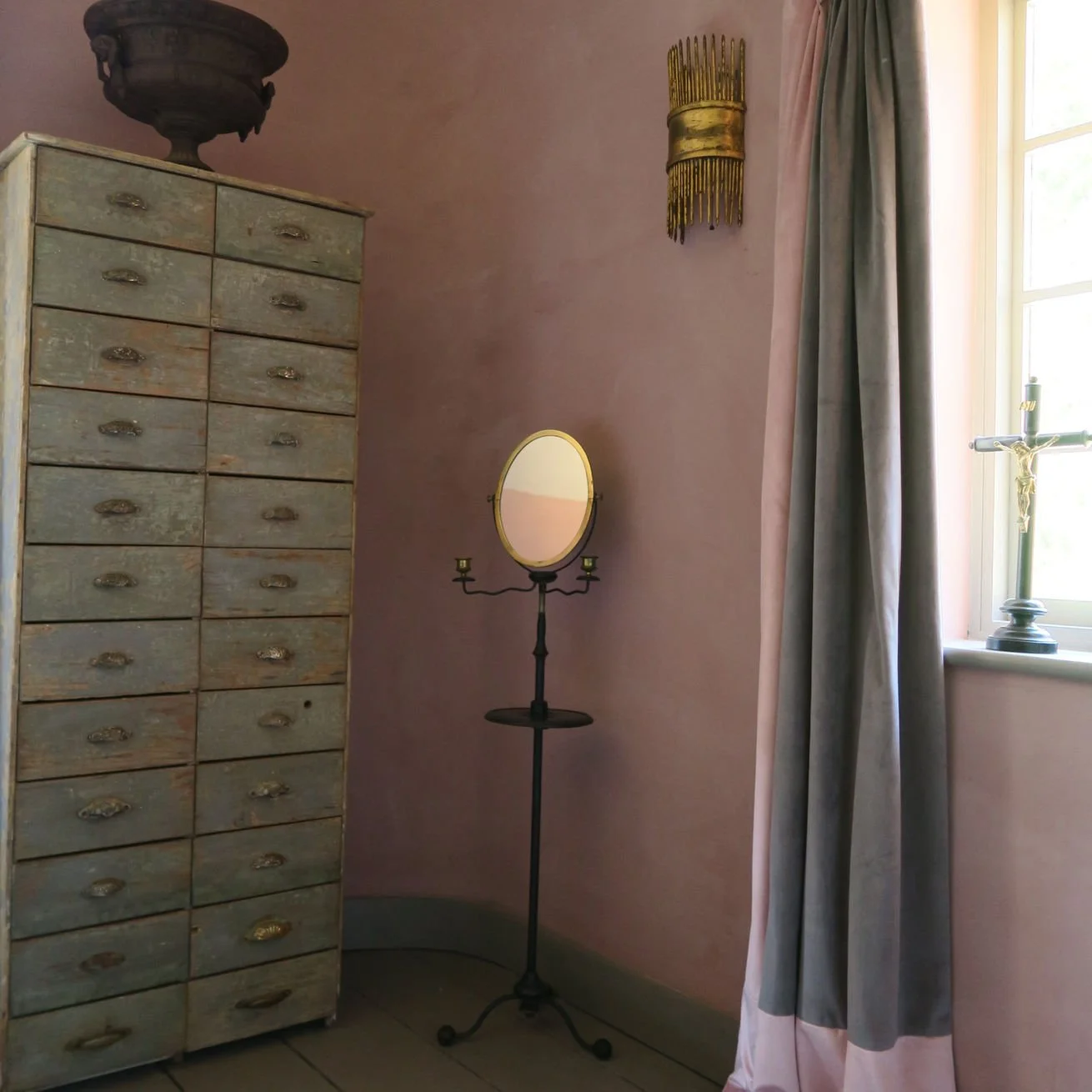 Vintage wooden drawer cabinet with numerous small drawers, a tall standing mirror with a gold frame, a candle holder on the mirror stand, a window with light-colored curtains, and a crucifix on the windowsill.