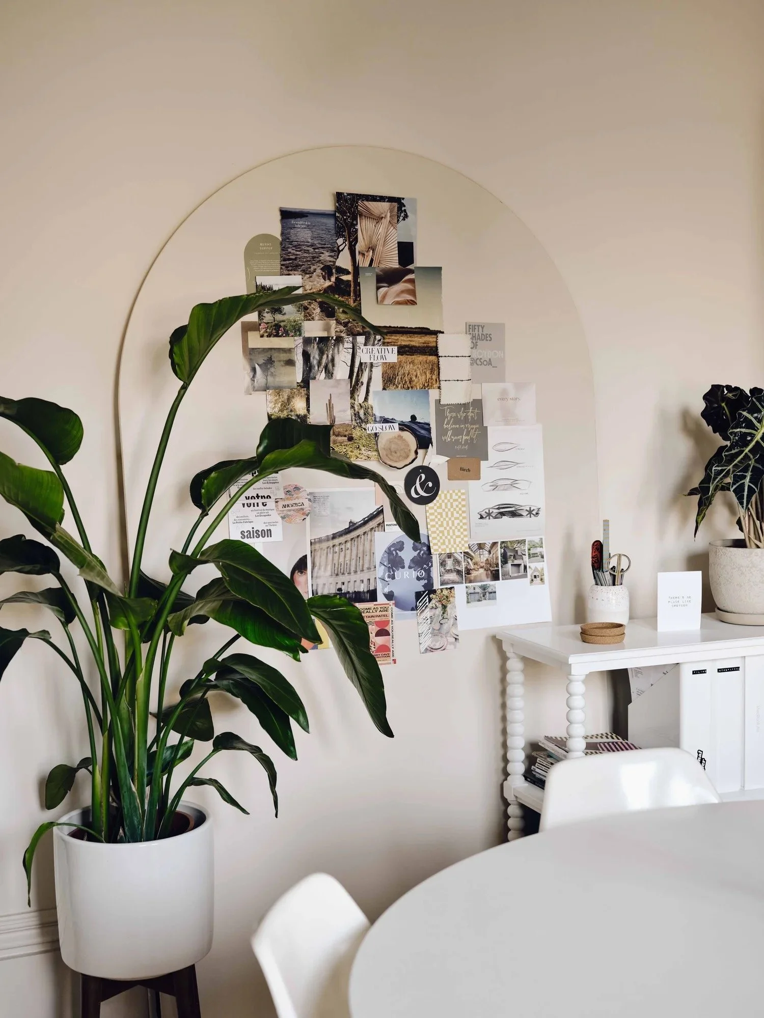 Indoor room with a white table, a white chair, and a white cabinet. There are two large potted plants and a bulletin board with photos and notes on the wall.