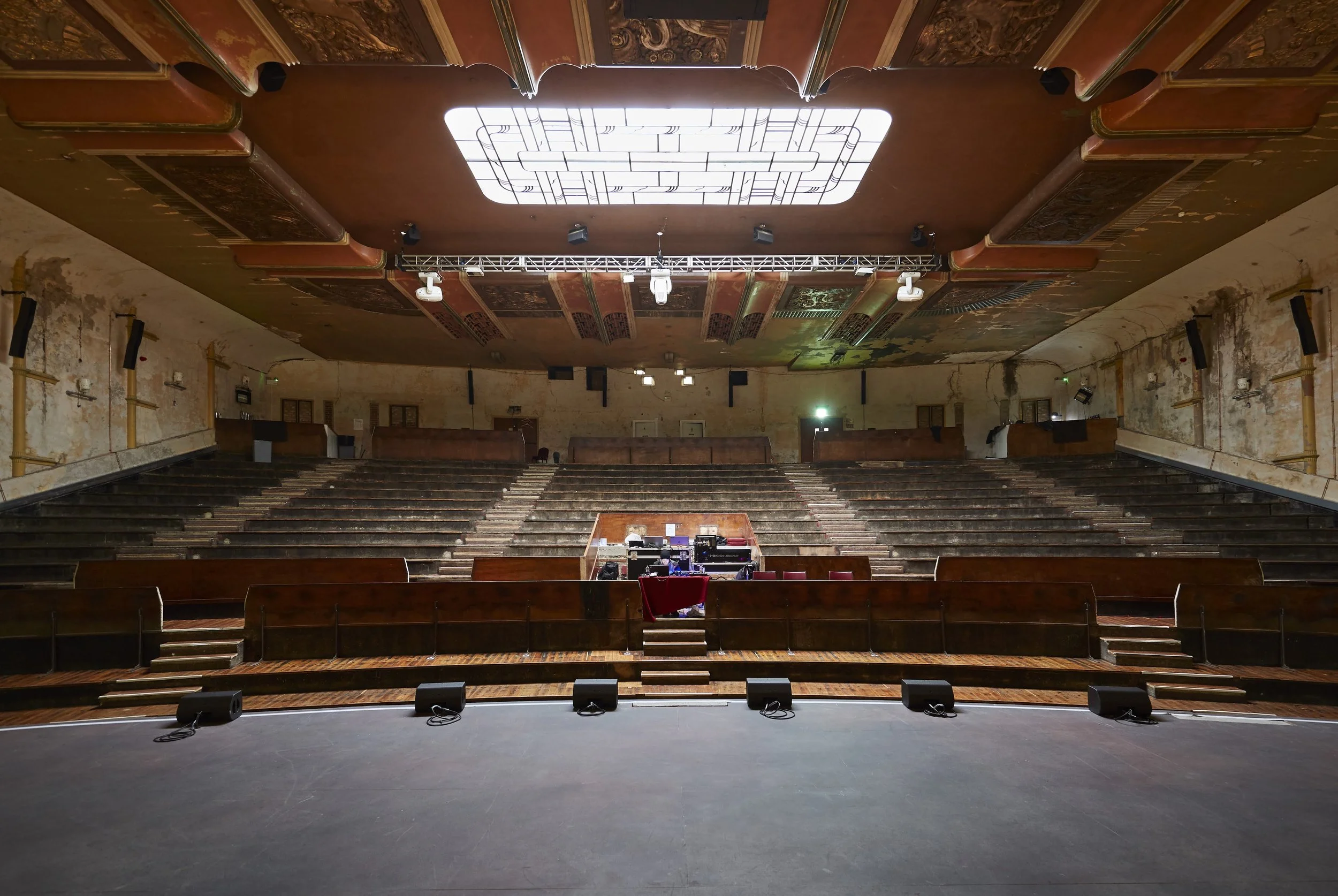 An empty theater or auditorium with a stage at the front, rows of wooden seats, and a large decorative ceiling light fixture.