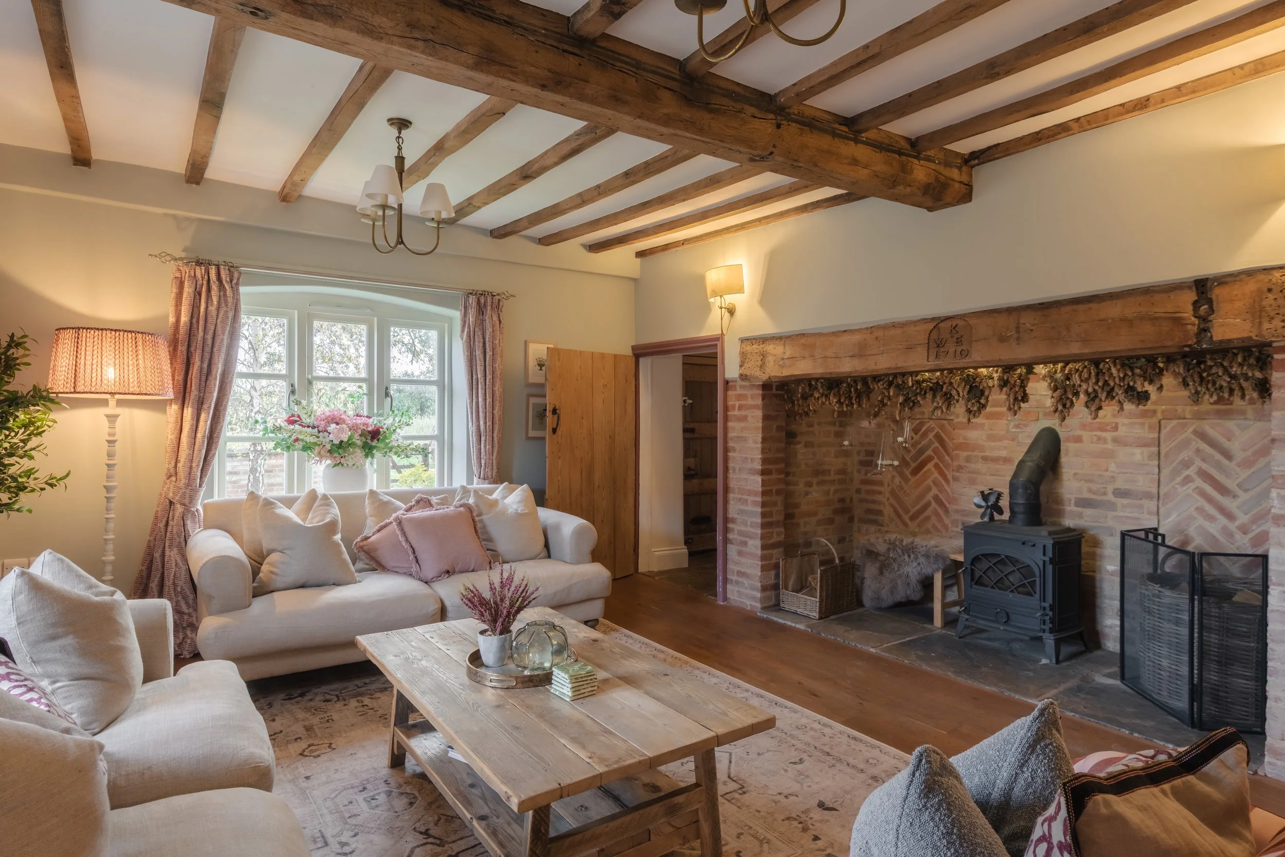 Cozy living room with beige sofas, pink cushions, pink curtains, a wooden coffee table, brick fireplace, wooden ceiling beams, floral centerpiece, and soft lighting.