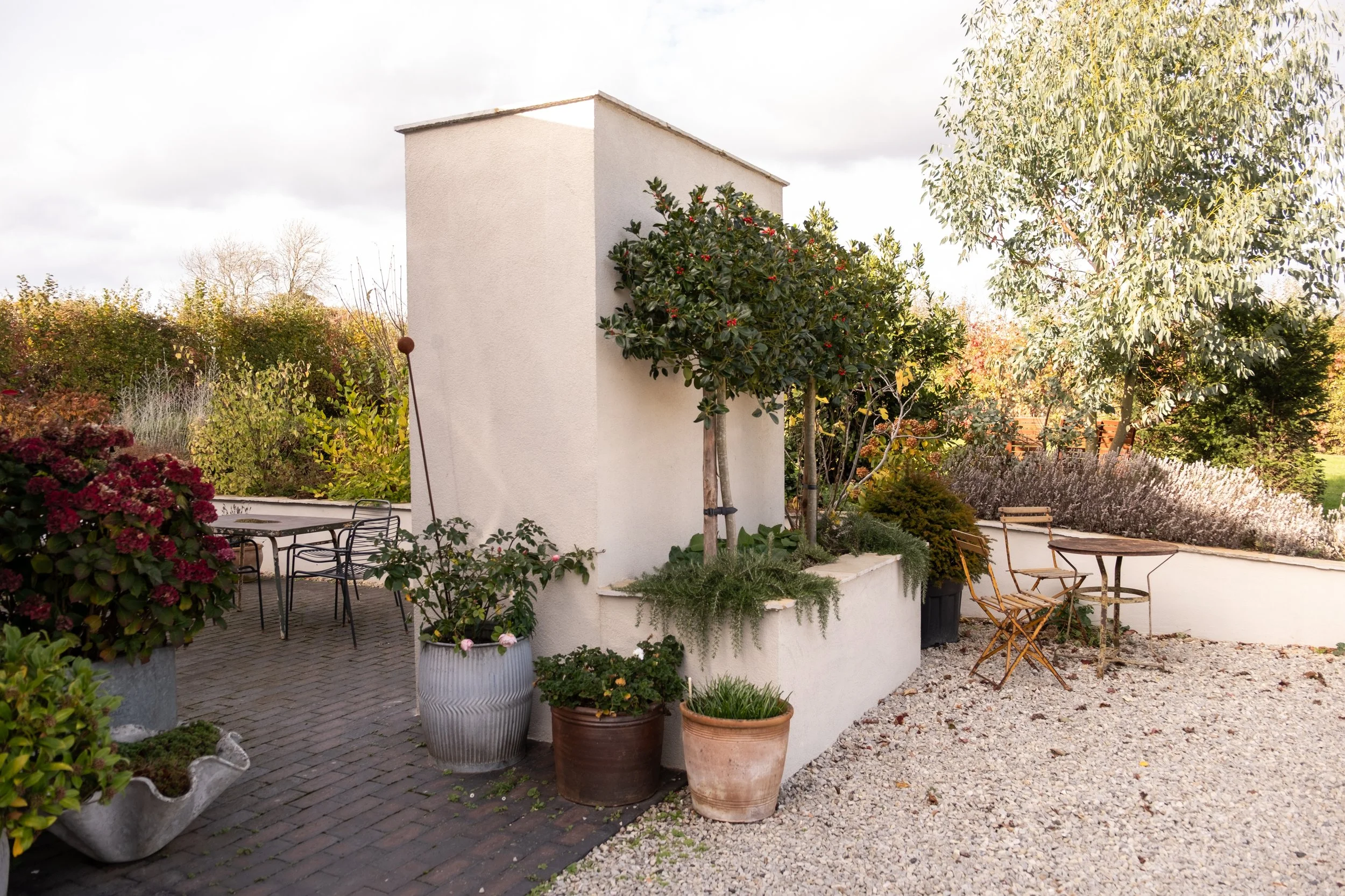 A small outdoor patio area with potted plants, a table, and chairs, surrounded by various trees and shrubs, with a white building in the background.