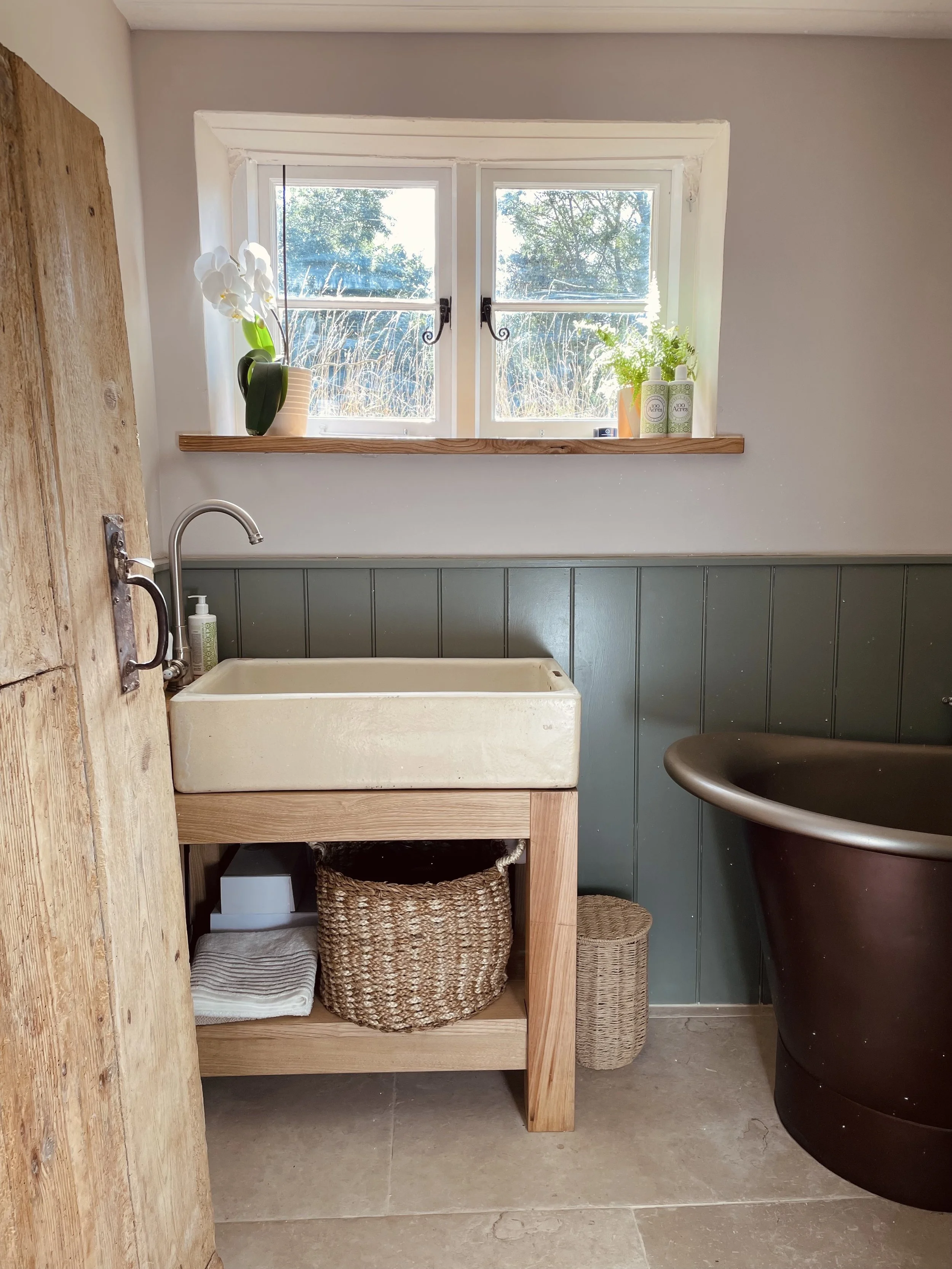 A bathroom with a white deep sink, a window with plants, a brown bathtub, a wicker basket, a towel, and a soap dispenser.