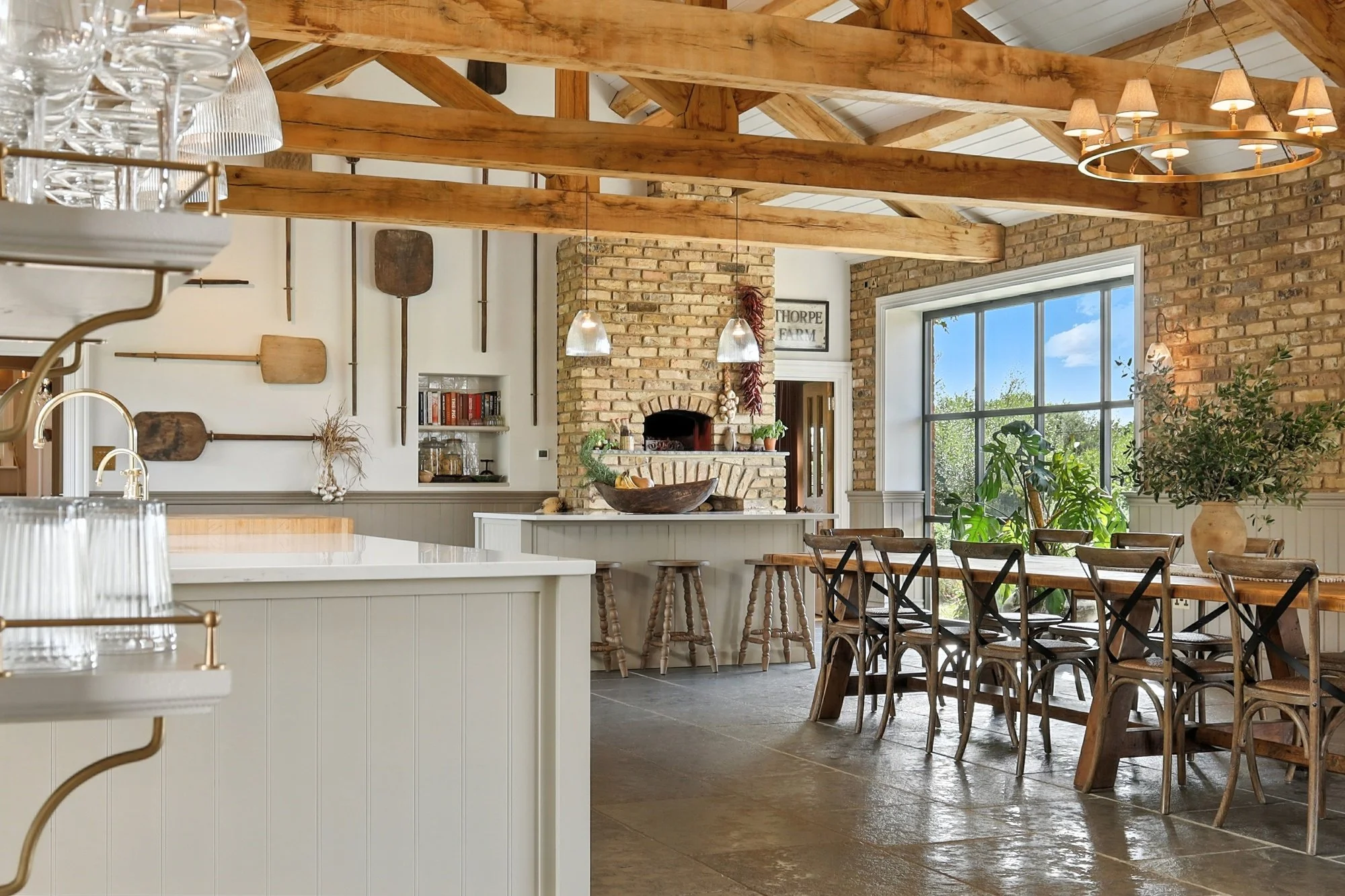 Interior view of a rustic-style dining area with exposed wooden beams, brick walls, a large window, and a wooden dining table with matching chairs. There is a fireplace with decor and a sign that says "Thorped" on the wall.