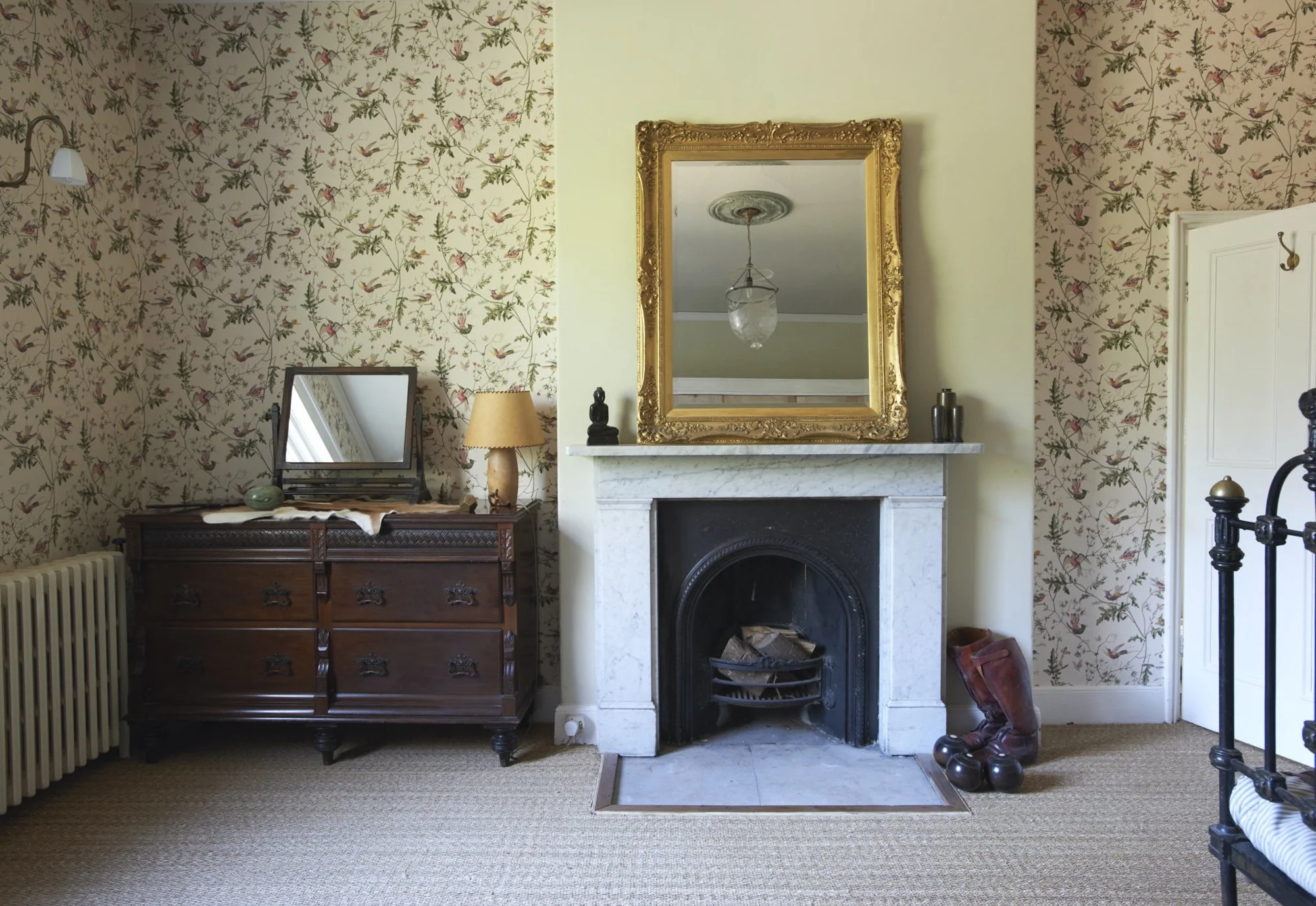 A vintage living room with floral wallpaper, a white marble fireplace with a black inner arch, topped with a large ornate gold mirror, a small black Buddha statue, and two black vases. To the left, there's a dark wooden dresser with a small mirror an