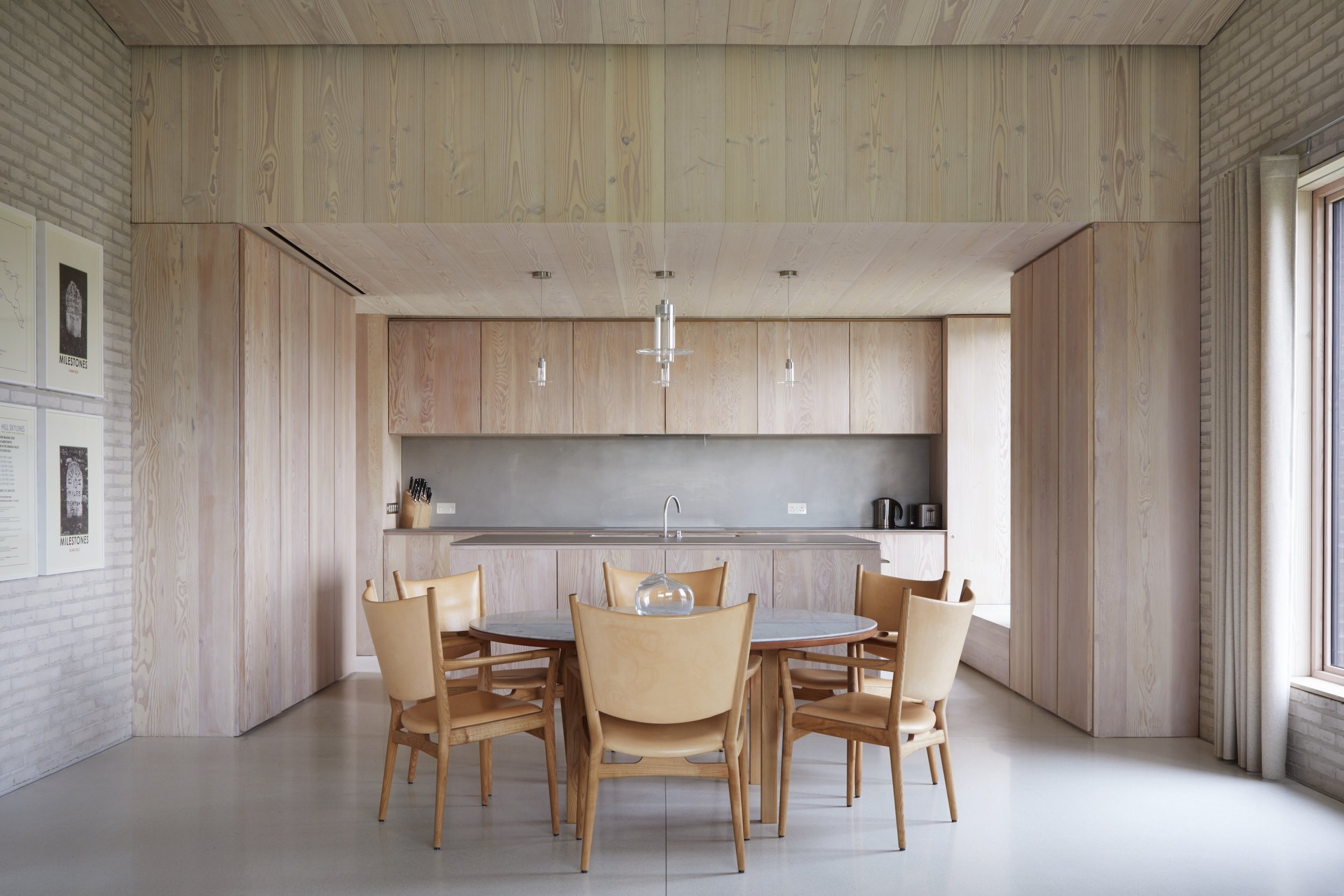 A modern kitchen and dining area with douglas fir cabinets, a round dining table, and eight coffee coloured chairs, with a large window on the right, ideal for lifestyle shoots and filming