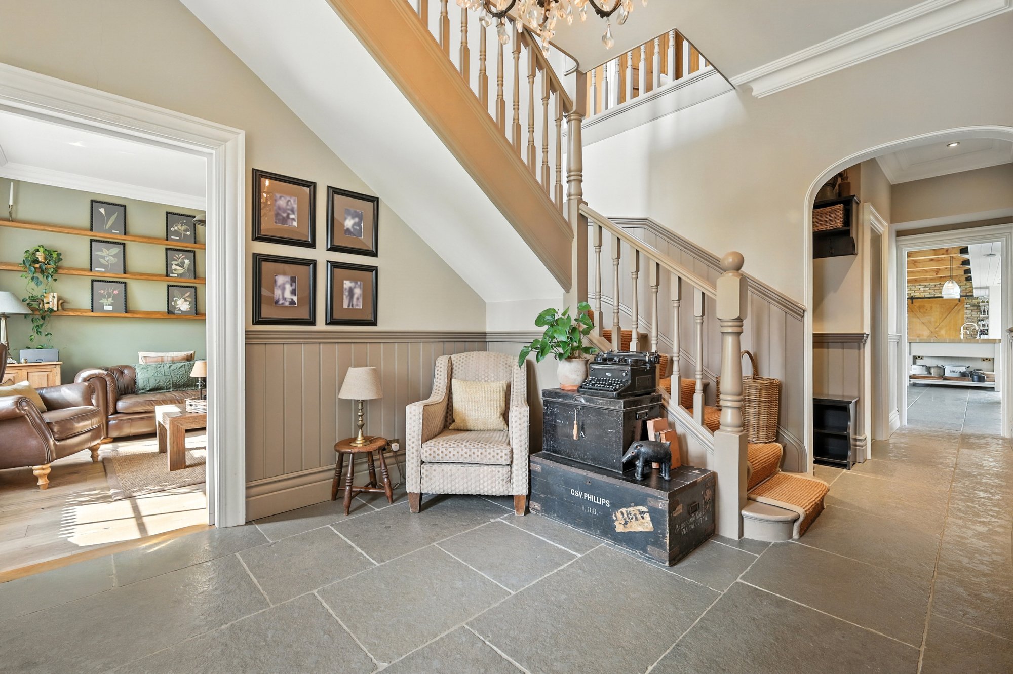 Living room and staircase with wooden railings, framed pictures on the wall, a chair with a potted plant, vintage crates, and a chandelier in a home interior.