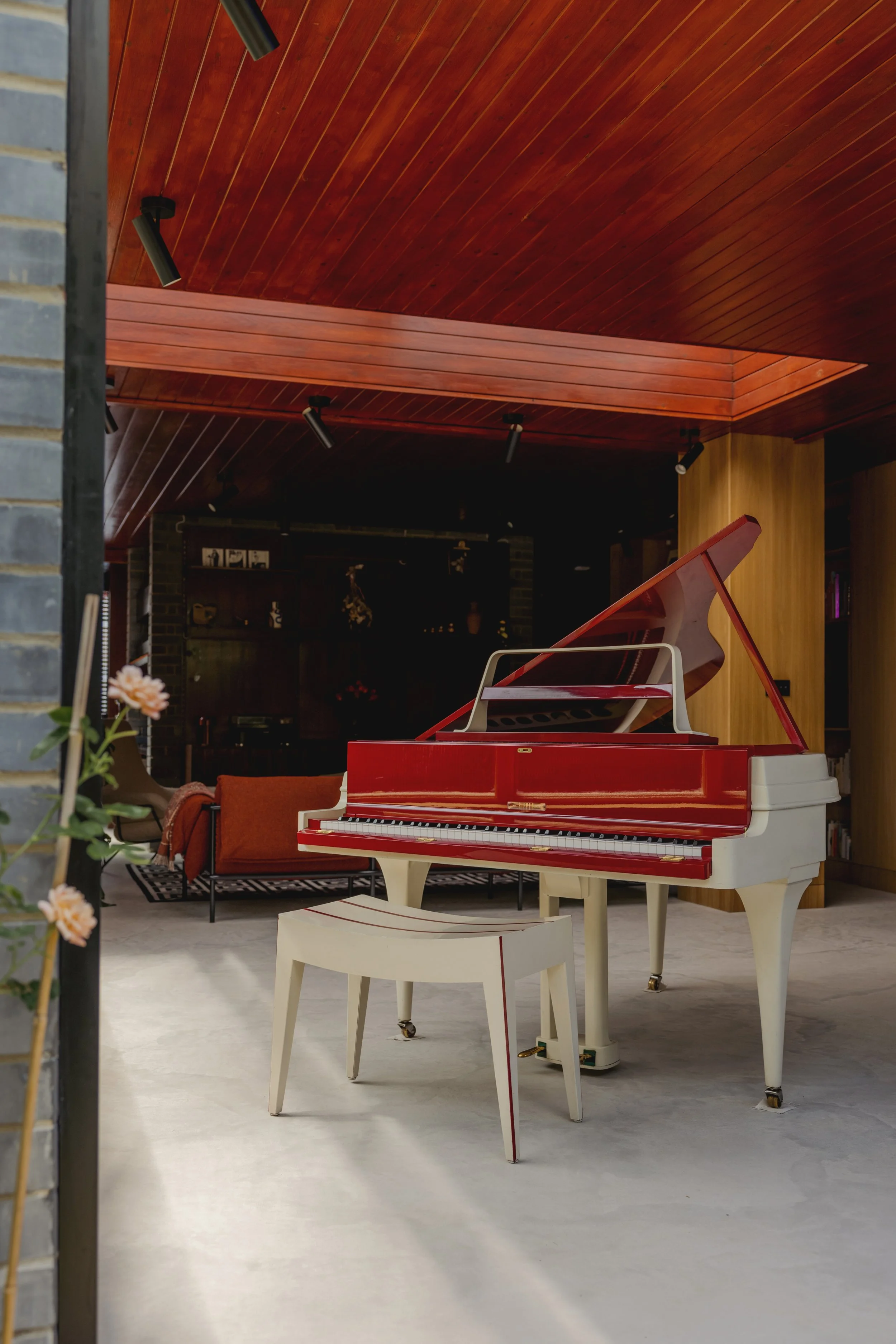 Red and white grand piano with matching bench in a modern living space with a wooden ceiling and a couch in the background.