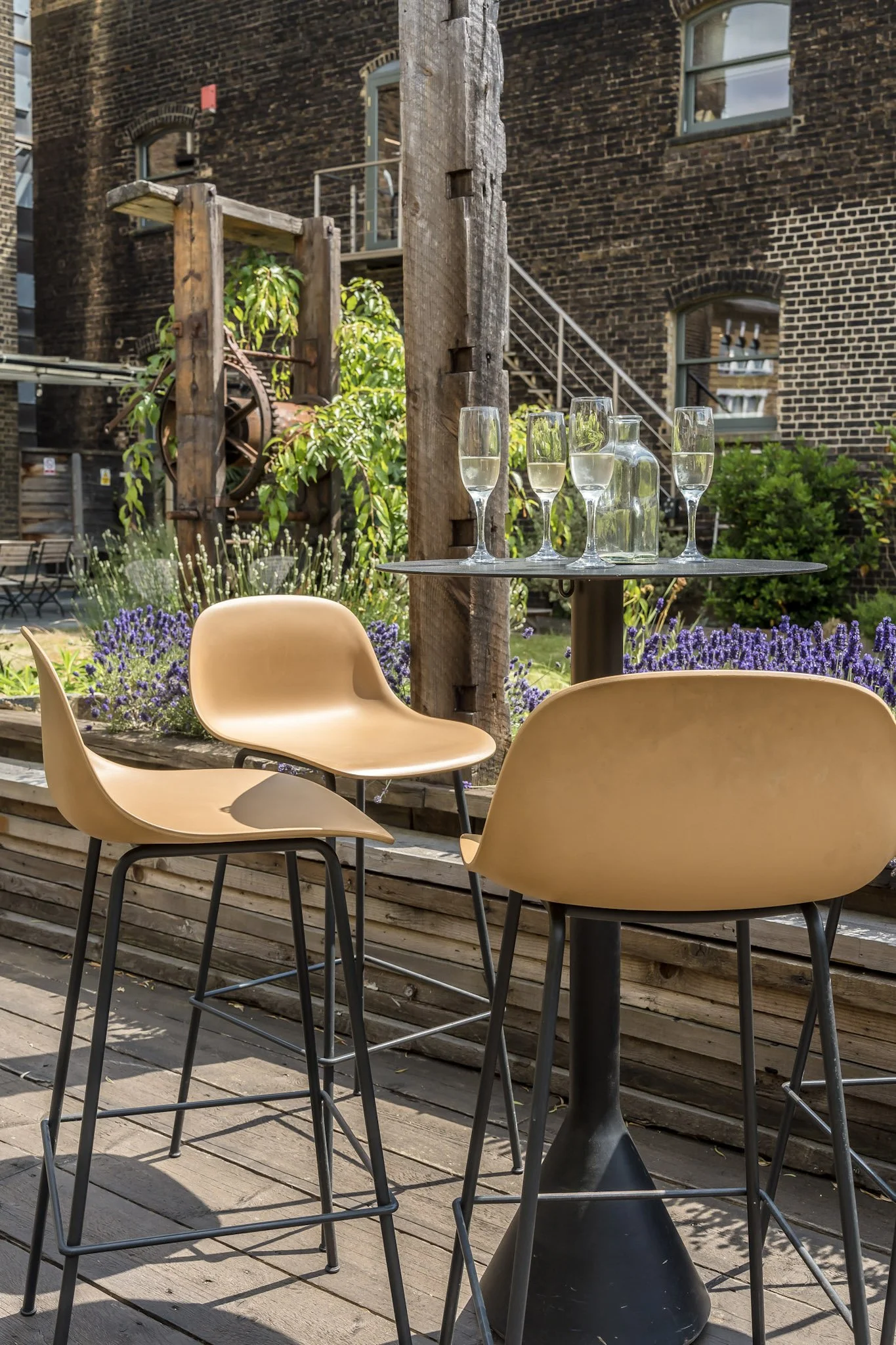 Outdoor patio table with three beige chairs and a black round table holding five champagne glasses and a glass pitcher of water, surrounded by purple flowers and wooden deck flooring with a brick building in the background.