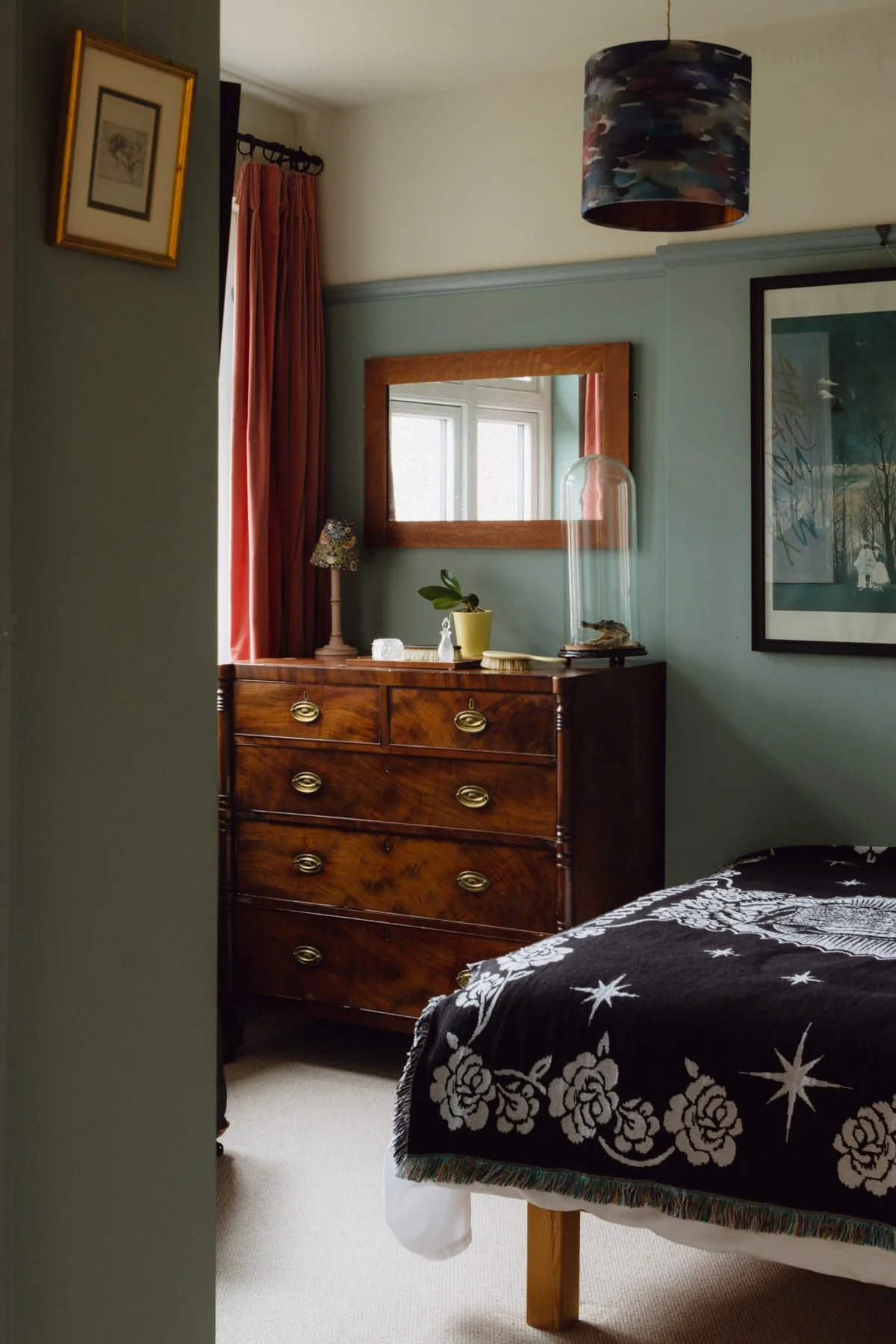 View of a bedroom corner with a wooden dresser, a mirror above it, a small lamp, and a window with red curtains. Part of a bed with a black and white floral blanket is visible.