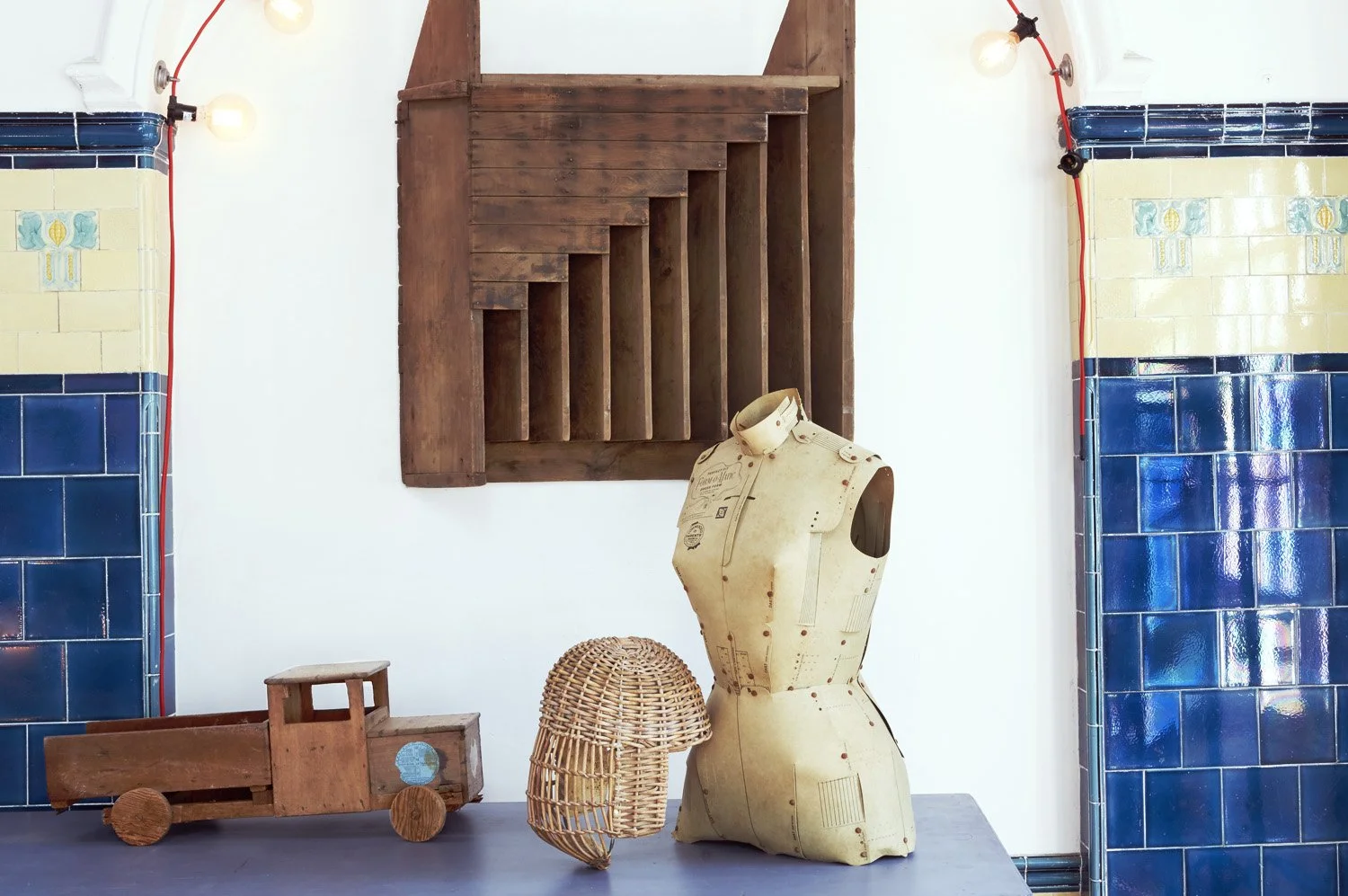 Interior of a room with blue and yellow tiled walls, a wooden ladder, a vintage dress form, a wicker hat, and a wooden toy truck.